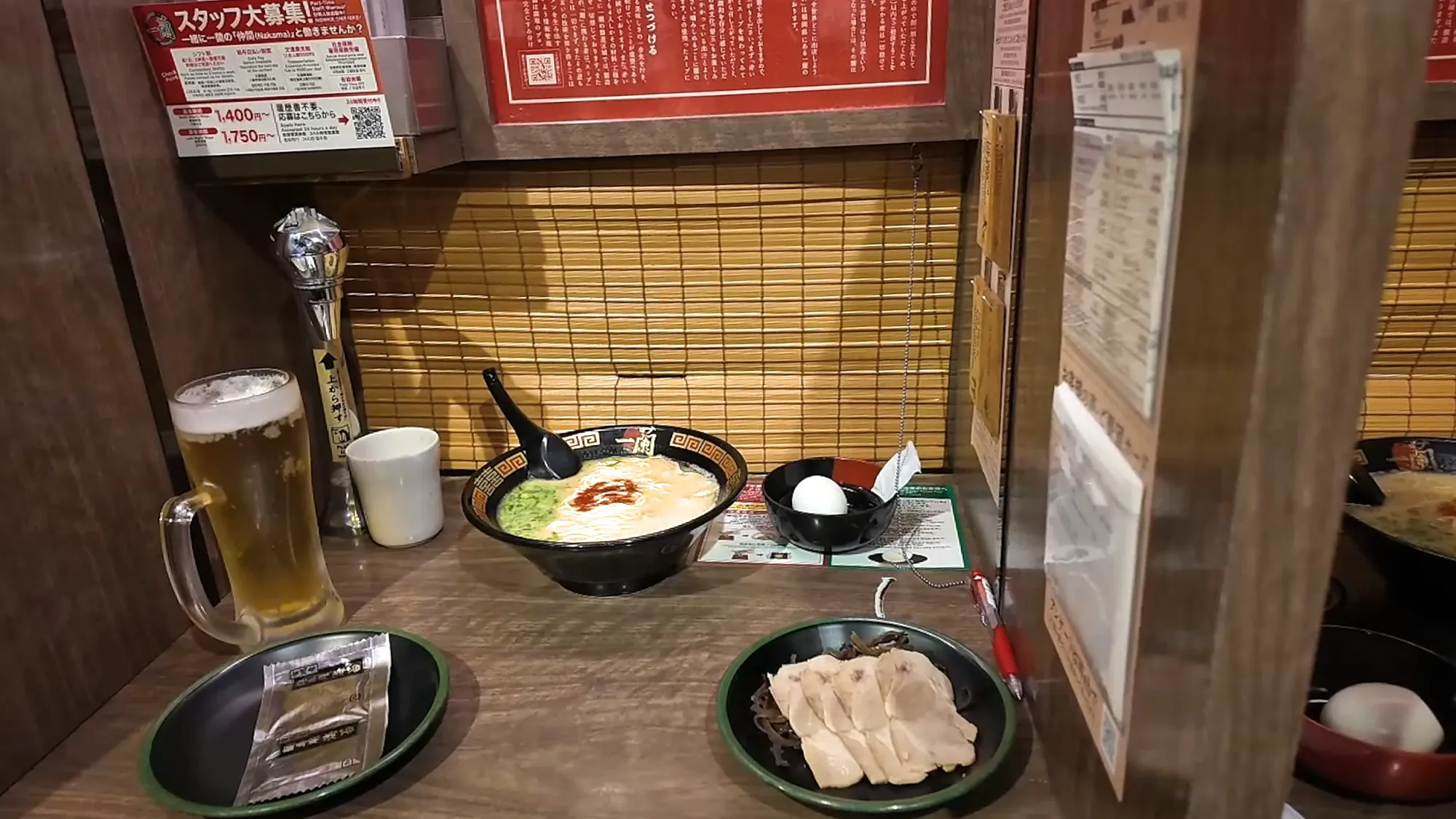 An Ichiran solo booth with ramen, a beer, a boiled egg, and extra pork, behind a bamboo blind