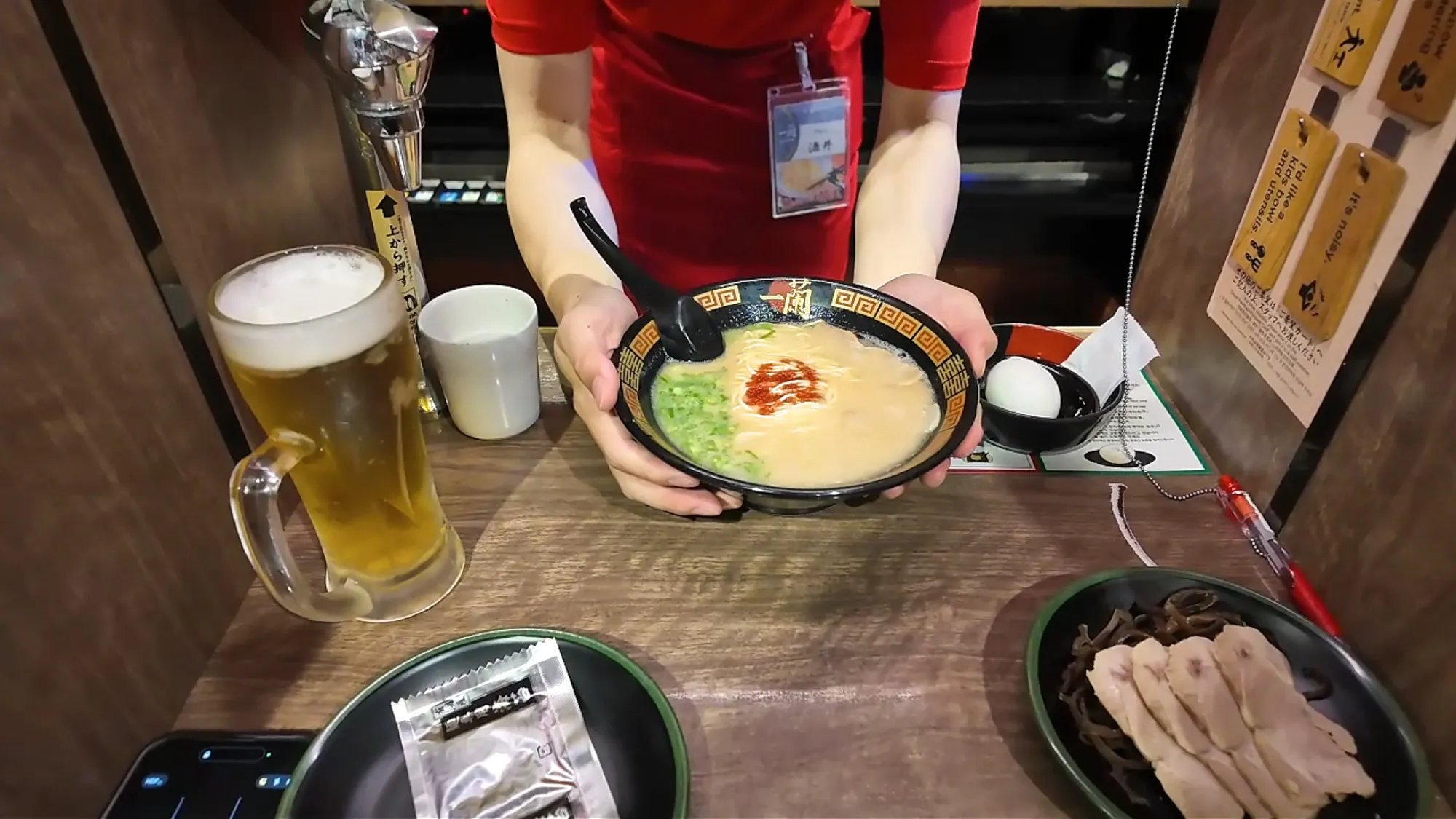 An Ichiran staff member setting a bowl of tonkotsu ramen down through the booth window, next to a draft beer, a salted egg, and a plate of chashu.