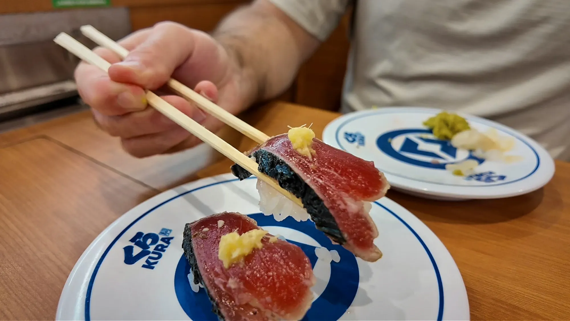 Lifting seared bonito nigiri with chopsticks over blue-and-white Kura Sushi plates in Tokyo
