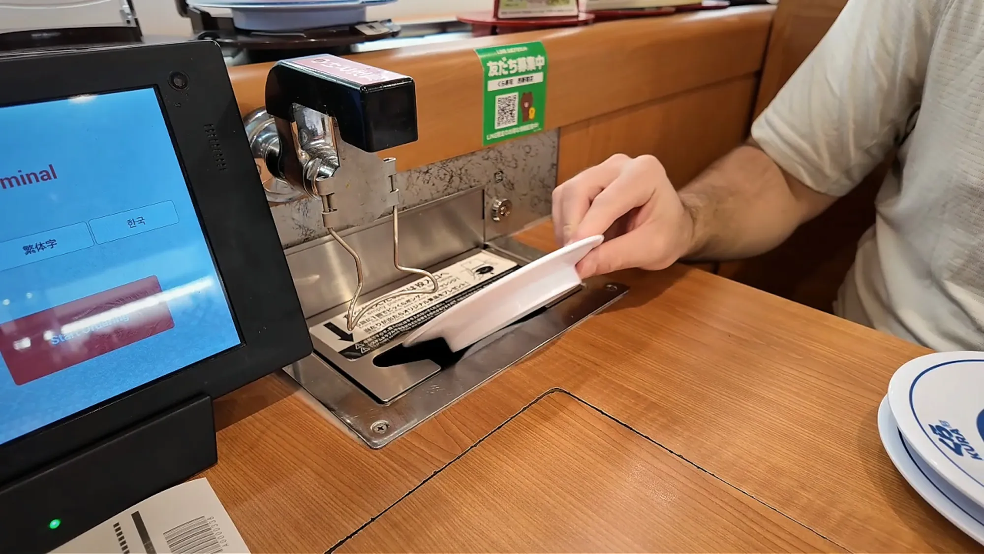 A hand sliding an empty plate into the plate return slot at a Kura Sushi table, beside the ordering tablet.