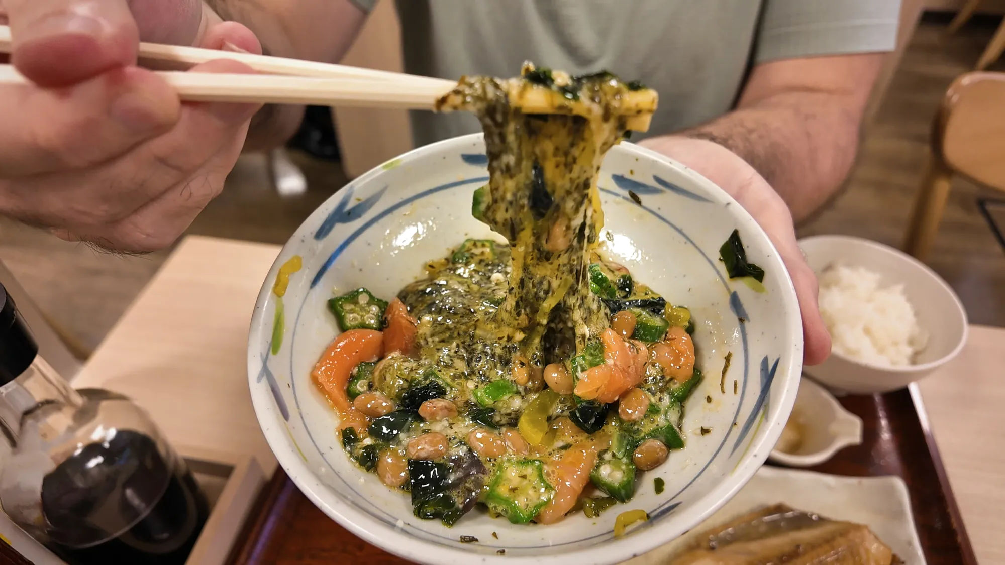 Lifting a stringy mix of natto, okra, seaweed, and fish from a neba-neba bowl with chopsticks at Yayoiken