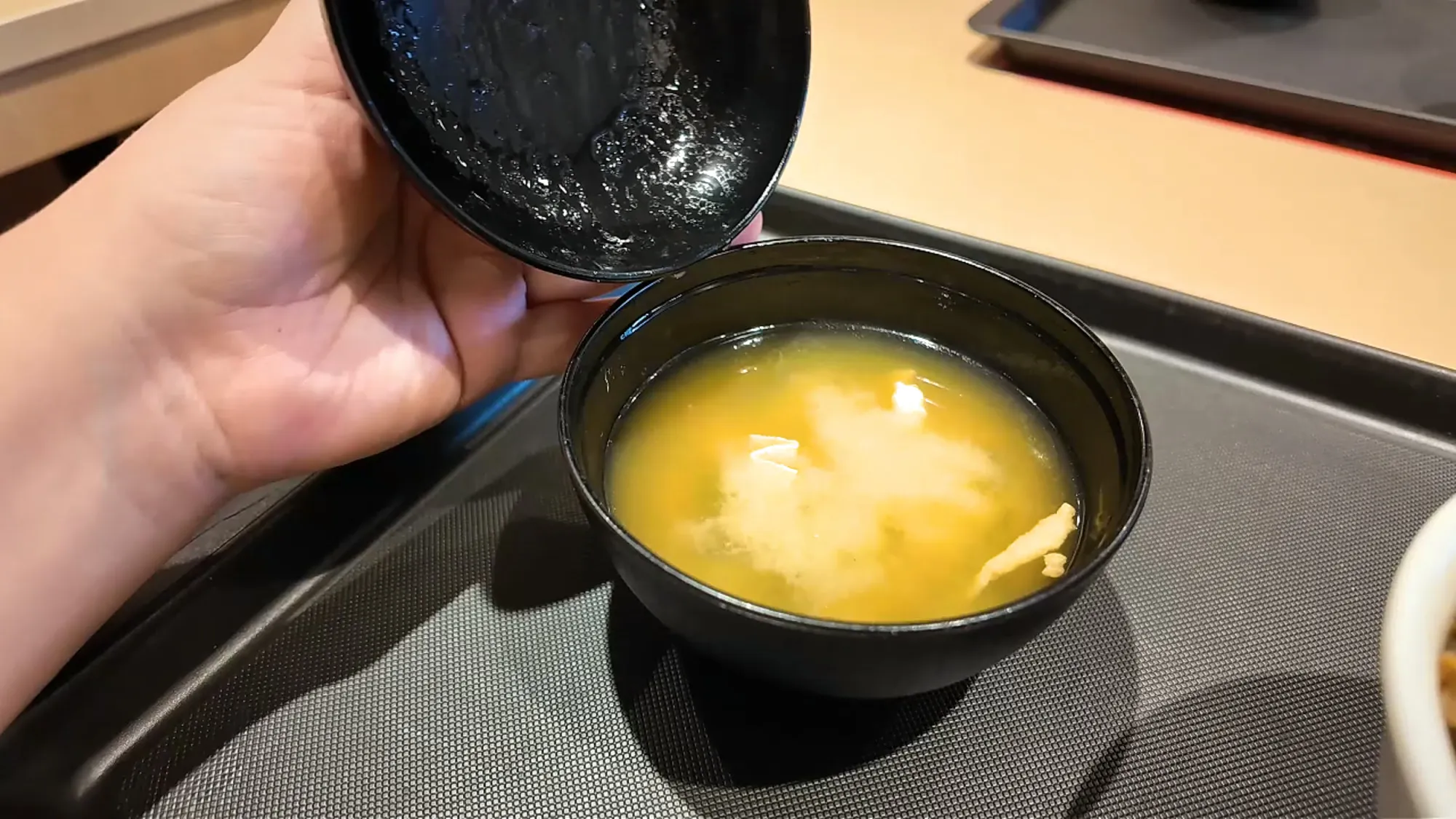 A hand lifting the lacquered lid off a bowl of miso soup with tofu at Yayoiken.