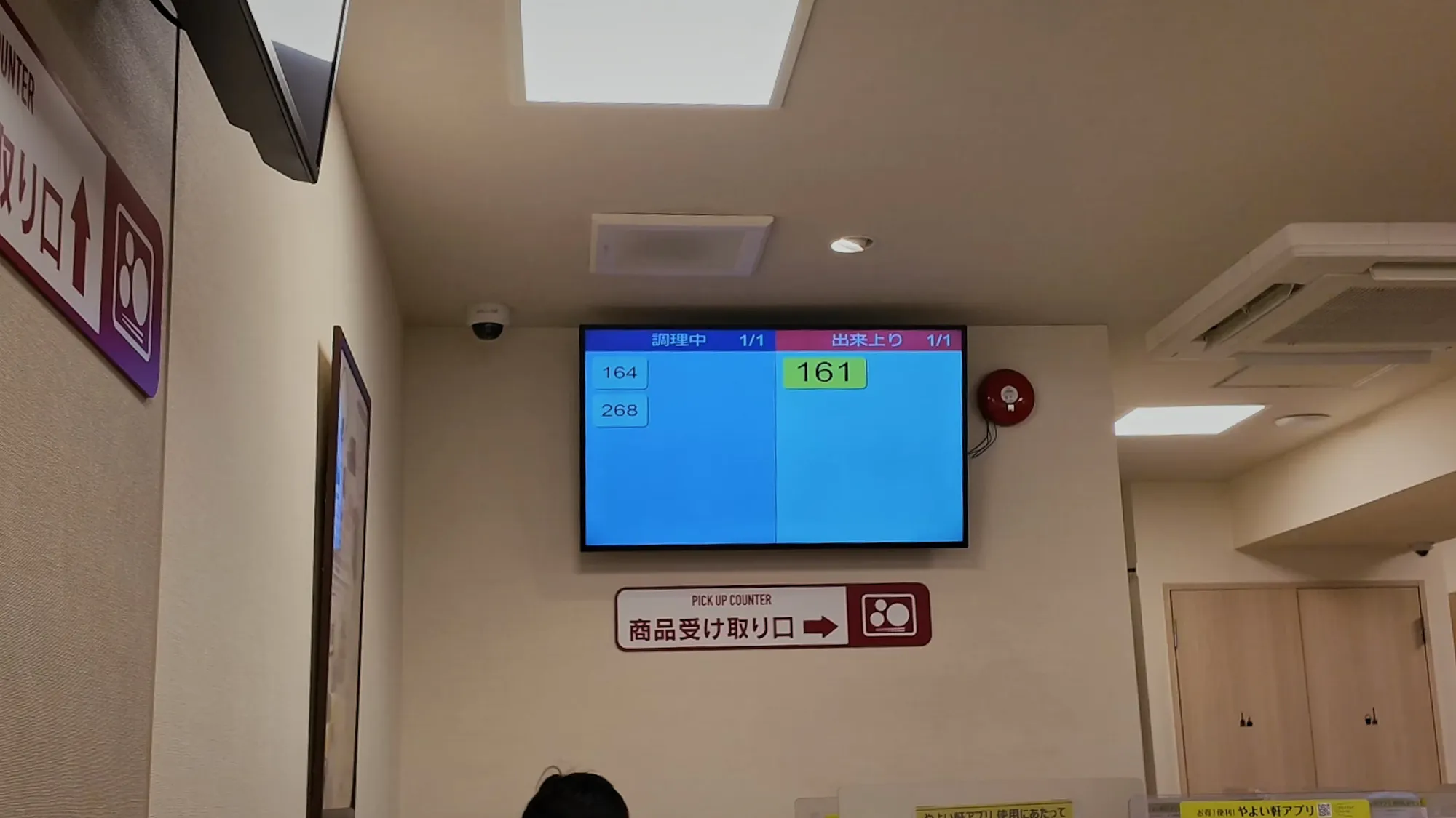 A screen in a Yayoiken restaurant in Tokyo showing order numbers being prepared and order number 161 ready, above a pickup counter sign.