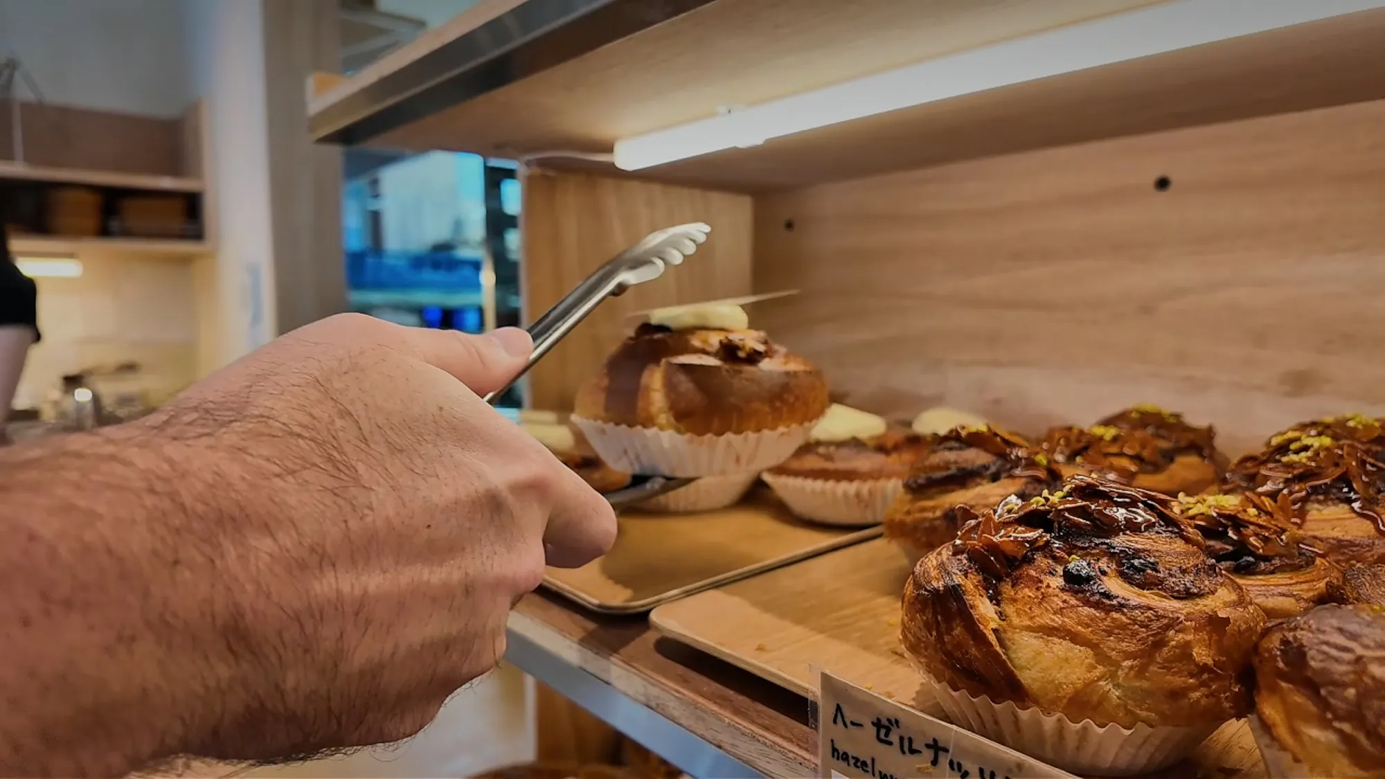 Using metal tongs to pick a fresh pastry off a wooden shelf inside a Japanese bakery in Tokyo