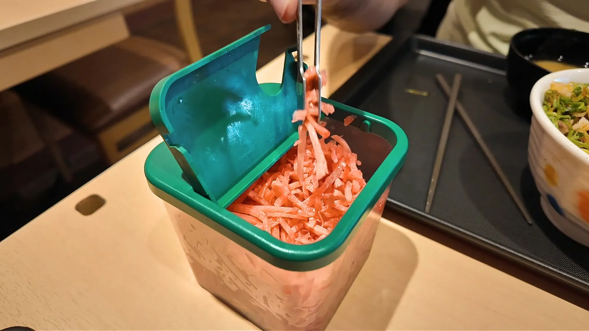 Tongs lifting pickled red ginger from a green tabletop container at Matsuya in Tokyo.