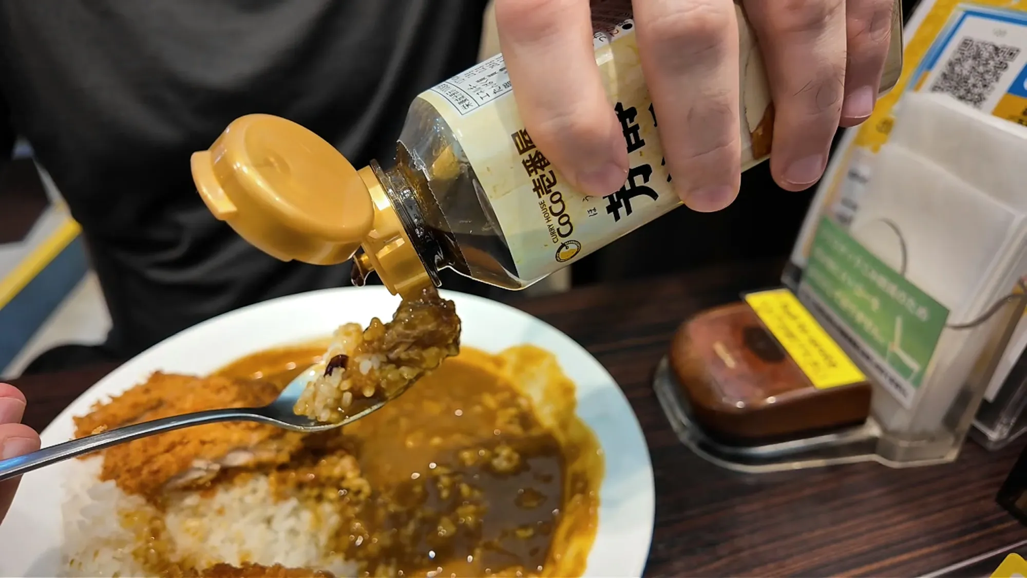 Pouring a thin stream of brown hojun sauce from the bottle onto a plate of katsu curry at CoCo Ichibanya.