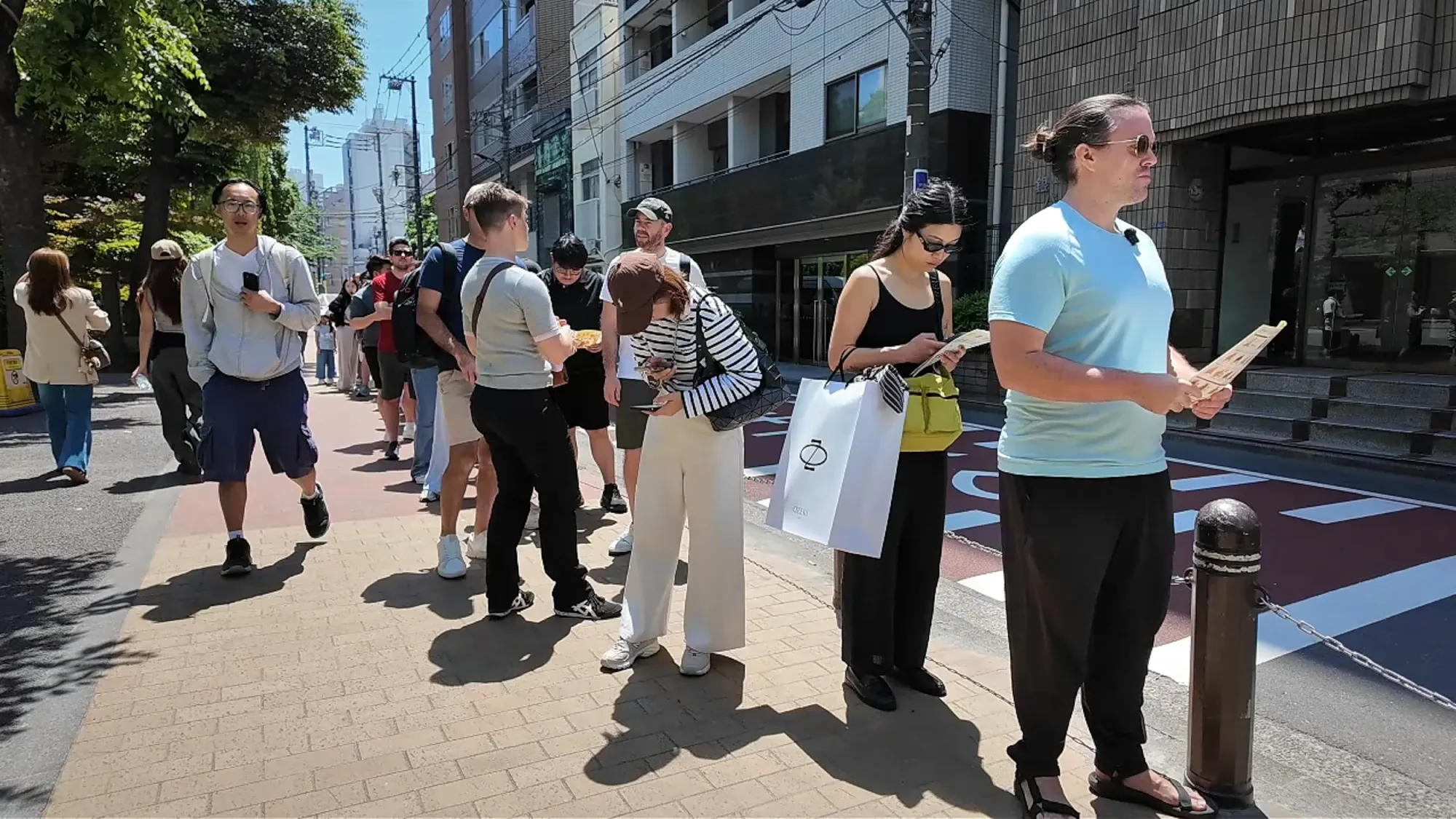 Cream holding a paper menu in a long line of people waiting on a sunny sidewalk outside Age.3 Ginza.