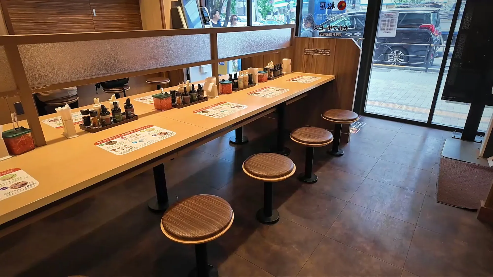 A long wooden counter with a row of round stools and condiments set out at each seat, inside a Tendon Tenya in Tokyo.