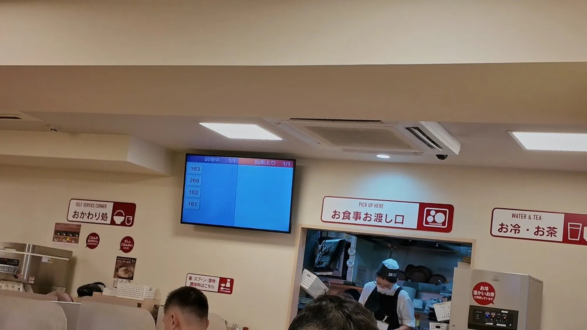 Inside a Yayoiken in Tokyo: the meal pickup counter, the self-service rice refill corner, and the water and tea station under red signs, with an order number screen above.