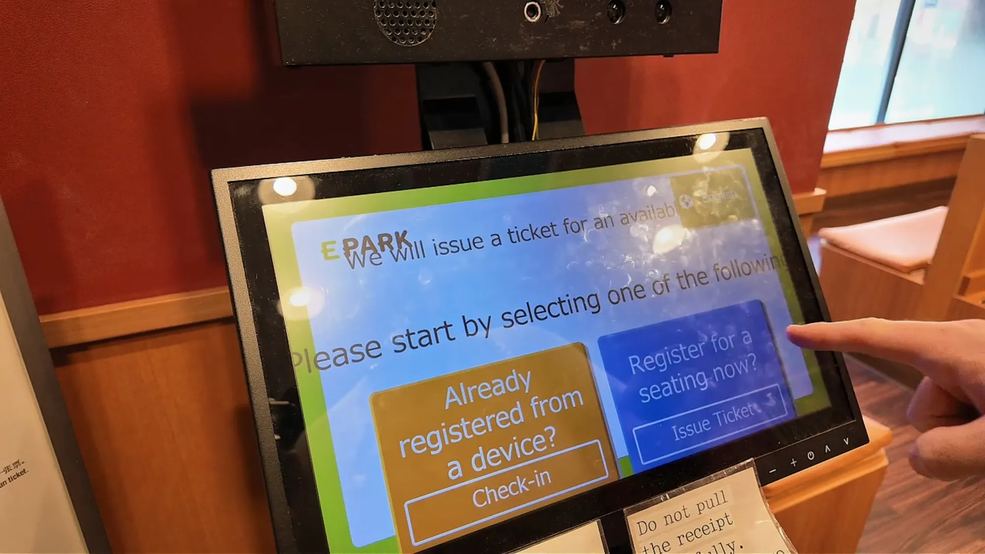 A finger tapping a restaurant check-in kiosk screen in Tokyo with English buttons for guests who already registered on a device.
