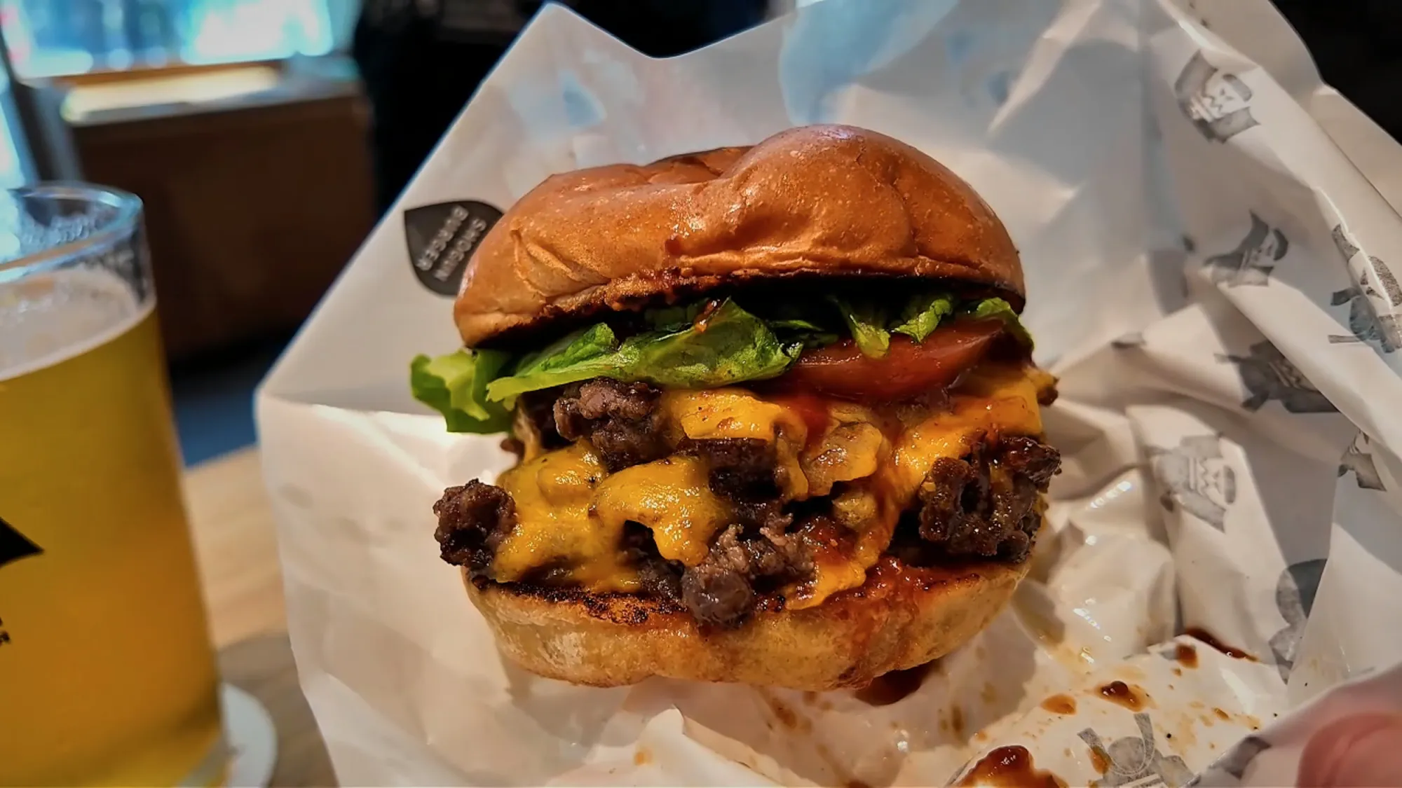 A double wagyu cheeseburger with melted cheddar, lettuce, and tomato in a paper wrap, with a draft beer behind it, at Shogun Burger in Shinjuku, Tokyo.