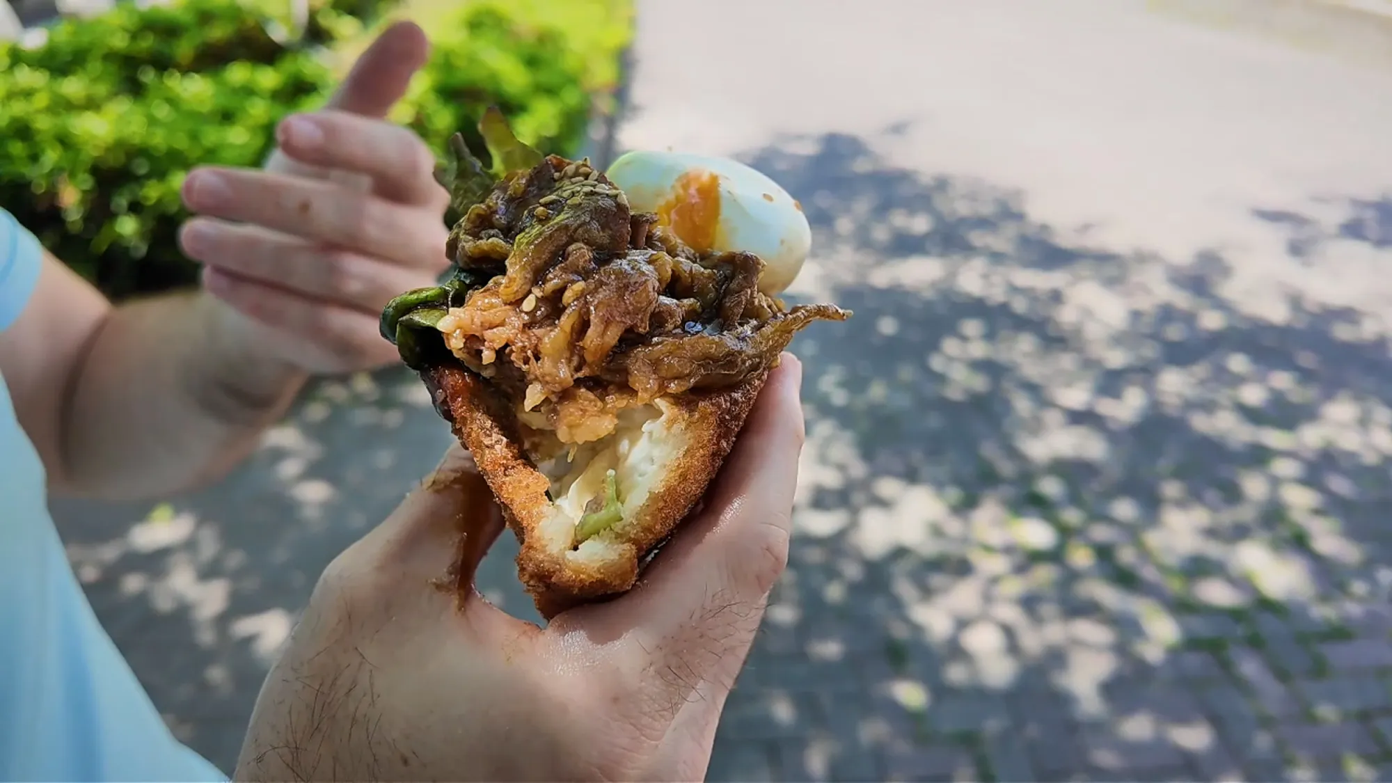 A fried sandwich stuffed with yakiniku beef short rib, potato salad, and a halved soft-boiled egg, held over a sidewalk in Ginza.