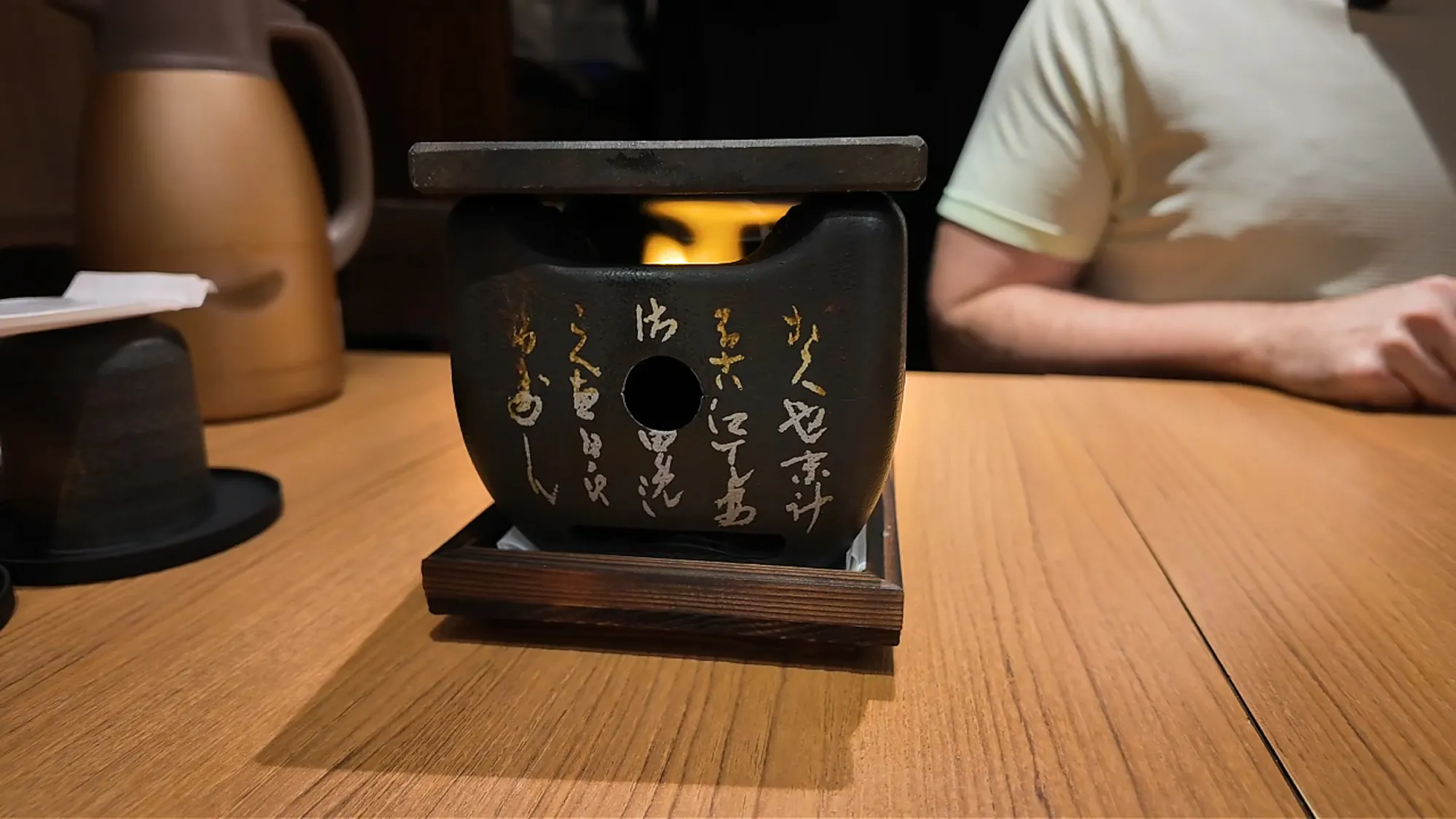 A small square hot stone on a black burner with a flame glowing underneath, on a wooden table at Gyukatsu Motomura.