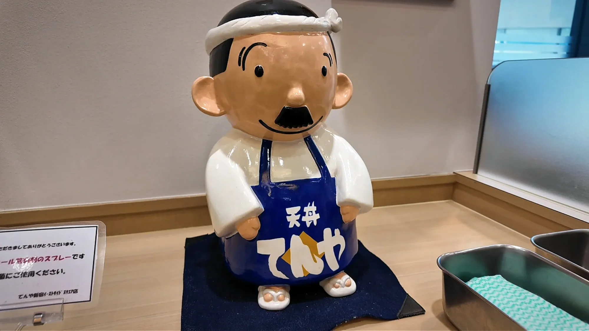 The Tendon Tenya mascot figure, a smiling cook in a headband and apron, inside a Tenya in Tokyo.