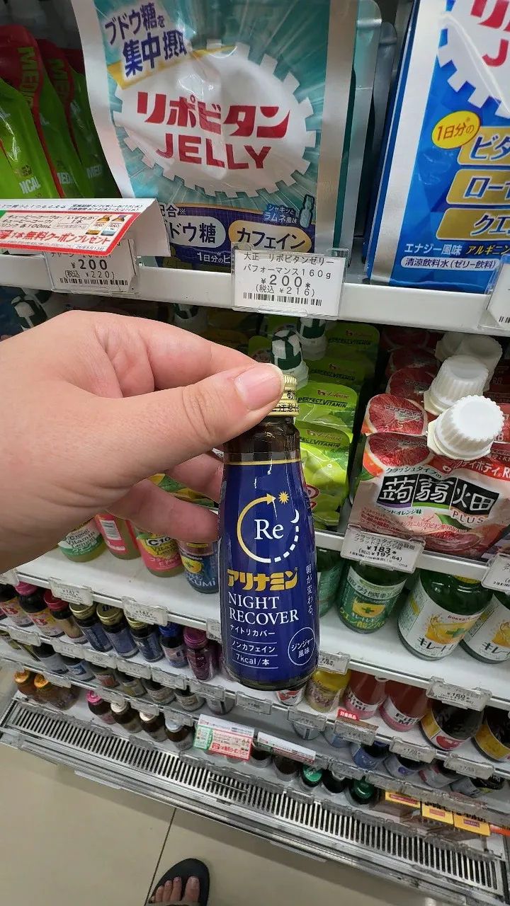 A hand holding a small navy blue bottle of Alinamin Night Recover, labeled caffeine-free and 7 kcal, at a 7-Eleven in Tokyo.