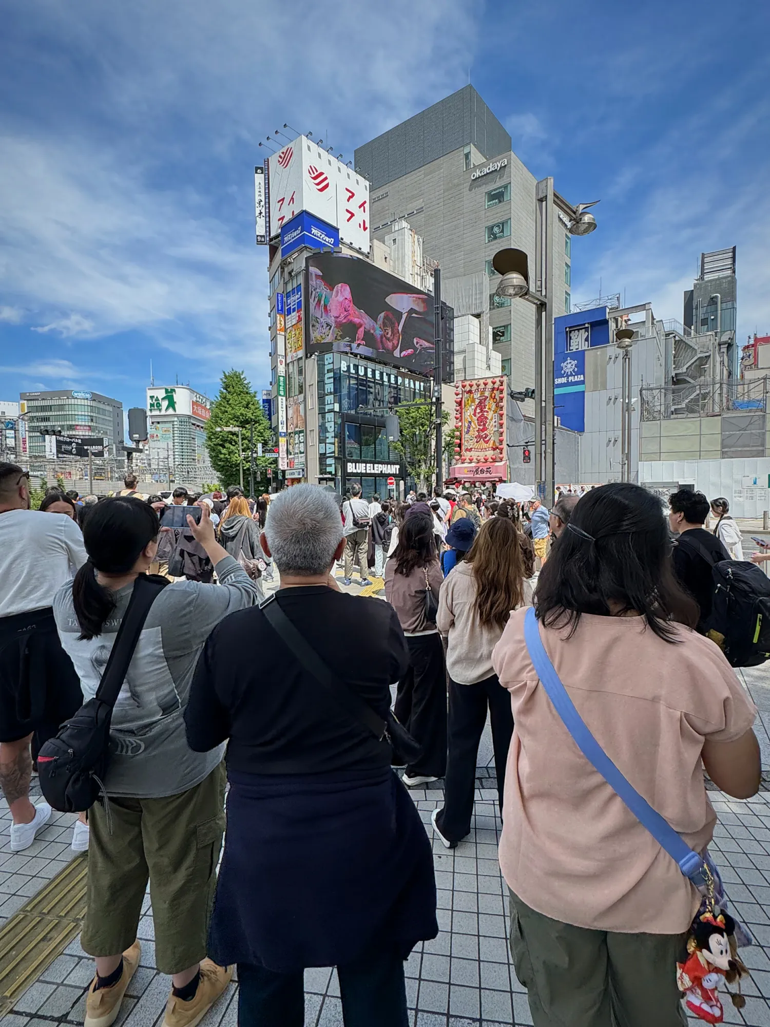 A crowd standing on the sidewalk outside Shinjuku Station's East Exit, looking up at the giant 3D billboard on the corner building.