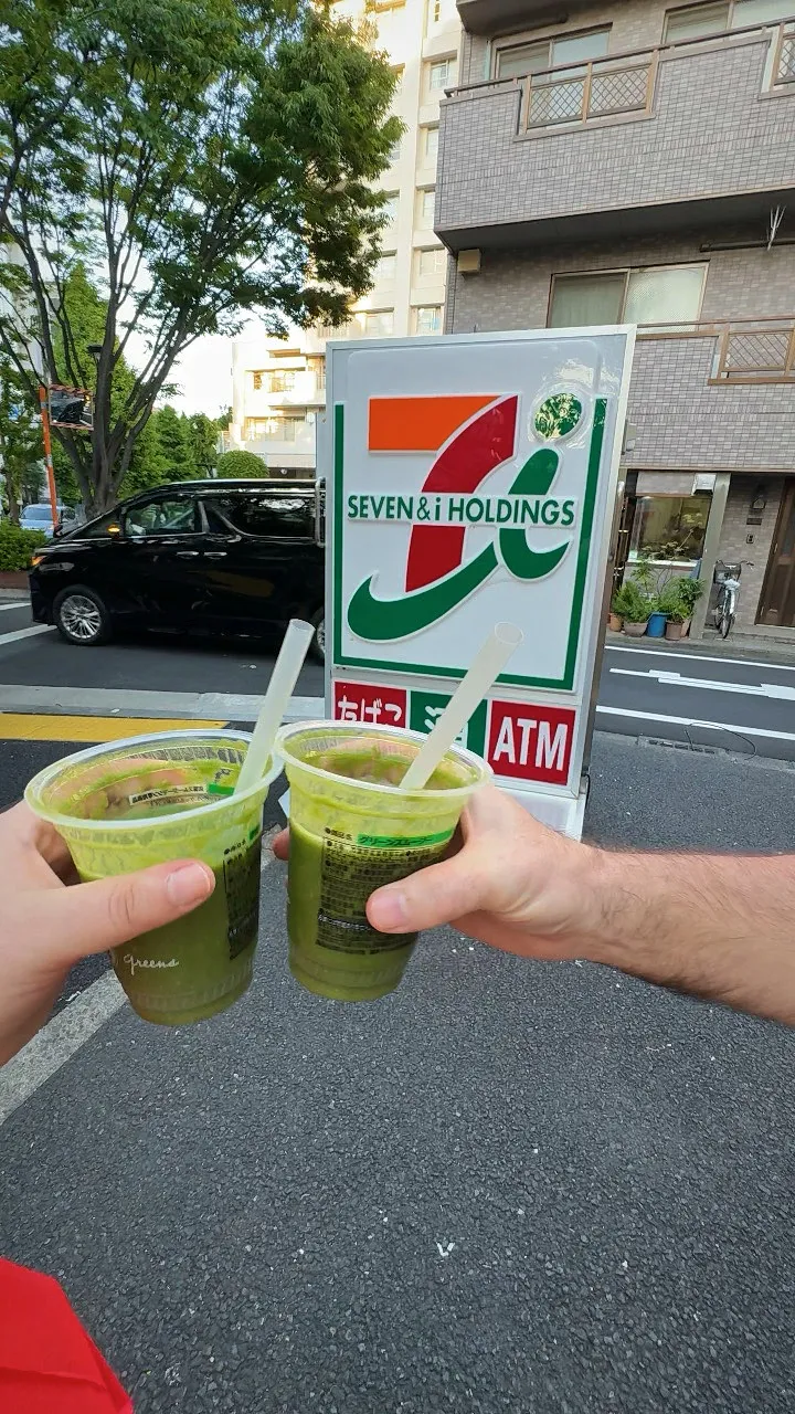 Two hands toasting with green smoothies in 7-Eleven cups in front of a Seven and i Holdings sign on a Tokyo street.