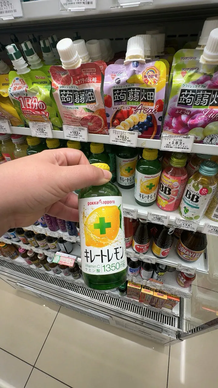 A hand holding a green Pokka Sapporo Kireto Lemon bottle labeled vitamin C and citric acid 1350 mg in front of a 7-Eleven drink shelf in Tokyo.