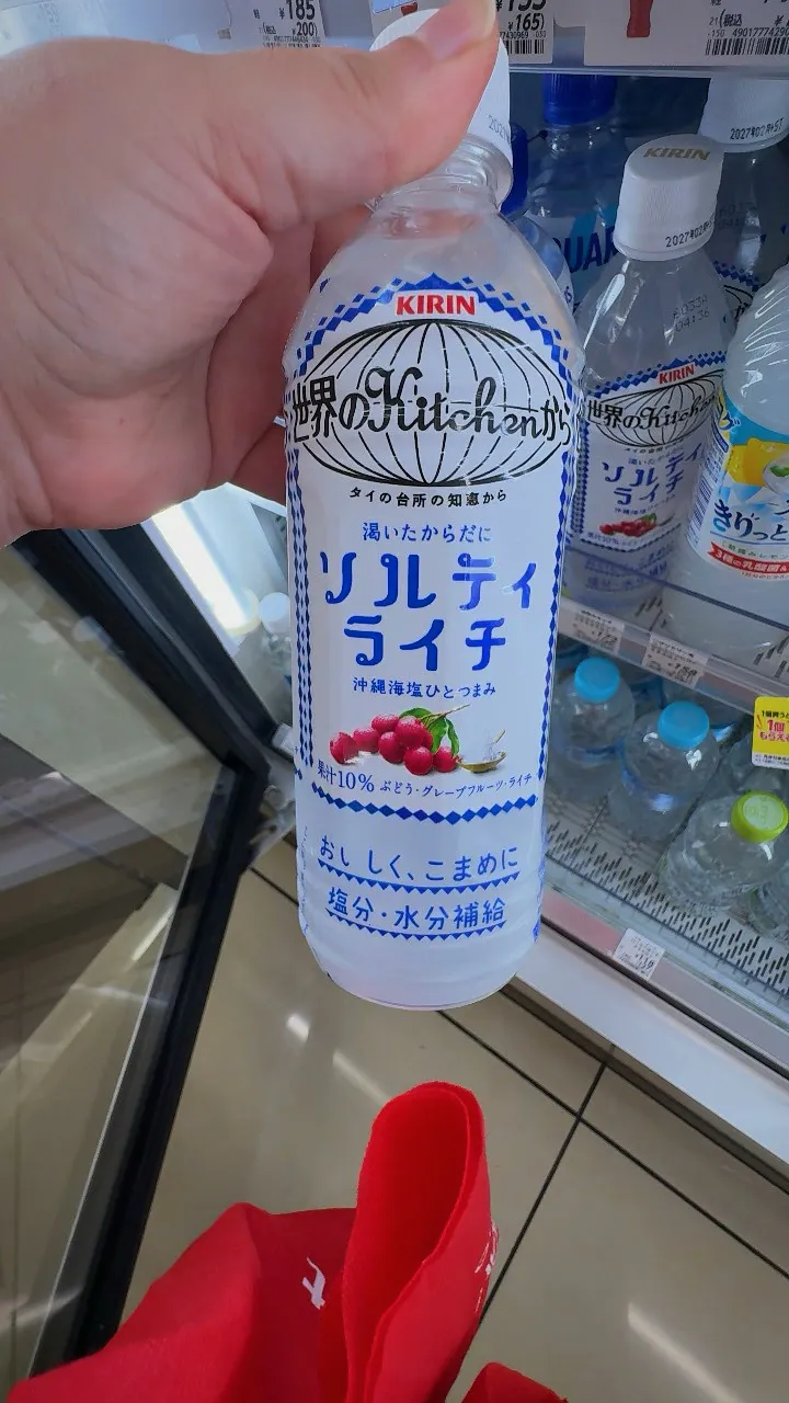 A hand holding a bottle of Kirin Salty Lychee drink in front of a convenience store drinks fridge in Japan.