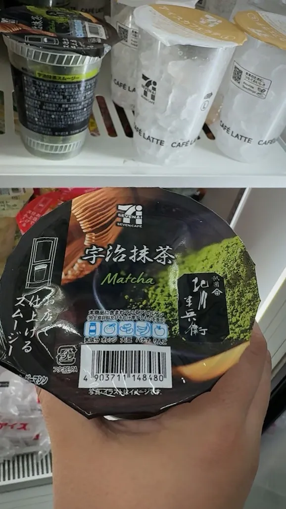 A hand holding a dark 7-Eleven Uji matcha smoothie cup with a green tea label in front of a store freezer in Tokyo.