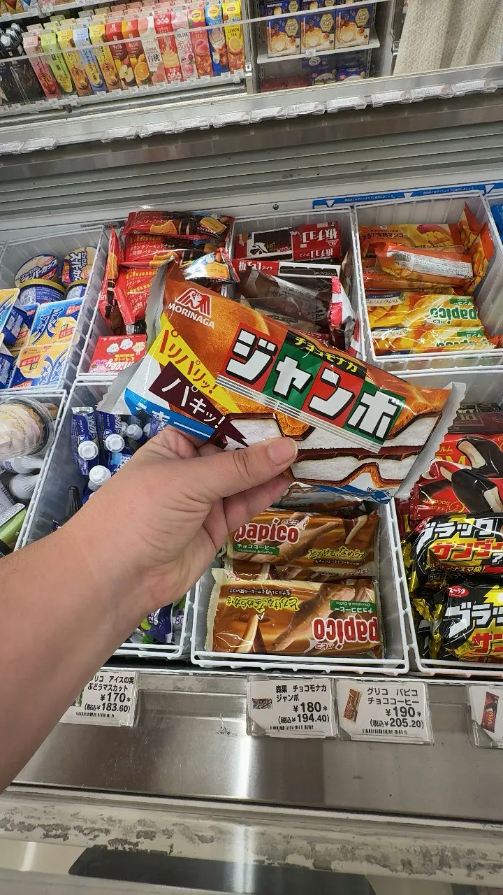 A hand holding a Morinaga Choco Monaka Jumbo ice cream sandwich above a 7-Eleven freezer in Tokyo, priced at 180 yen.