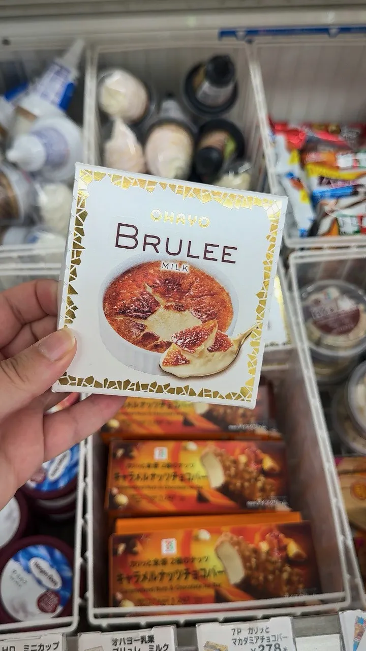A hand holding a box of Ohayo BRULEE crème brûlée ice cream above a 7-Eleven freezer in Tokyo.