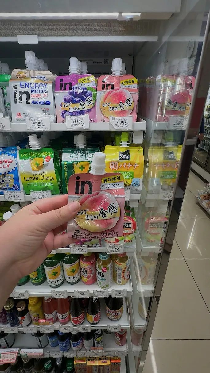 A hand holding a pink Morinaga in Jelly peach pouch with a dietary fiber and multivitamin label in a 7-Eleven fridge in Tokyo.