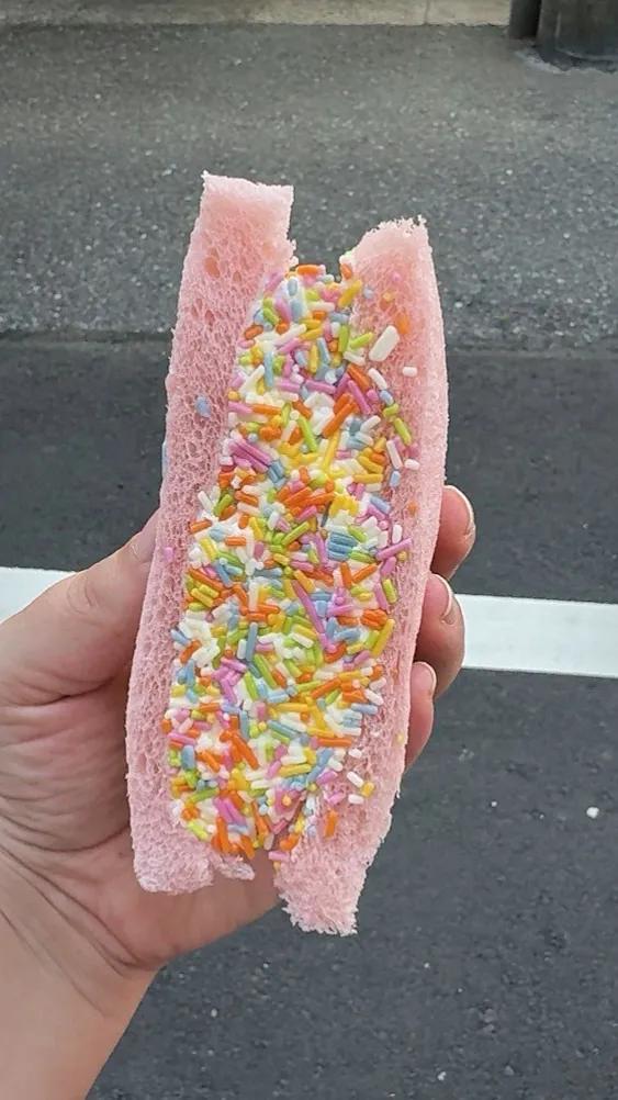 A hand holding a pink bread sandwich filled with whipped cream and rainbow sprinkles on a Tokyo street.