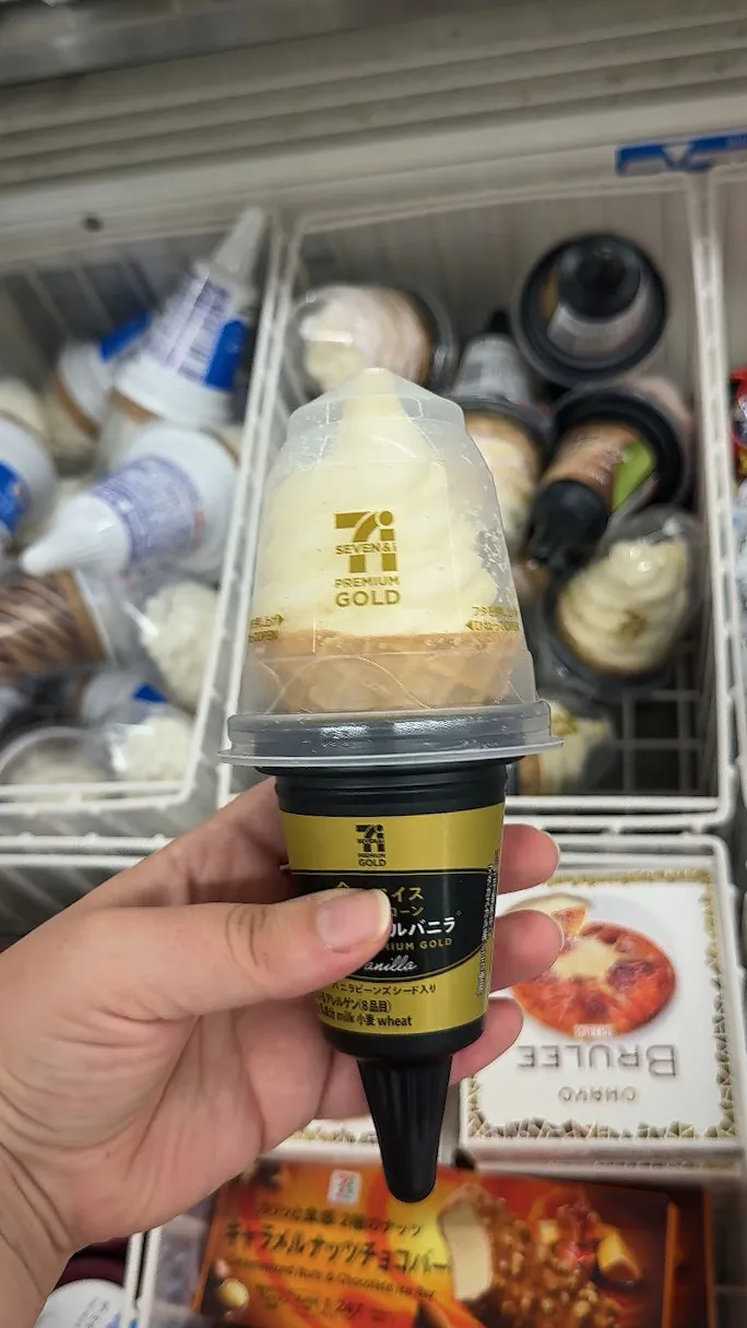 A hand holding a Seven Premium Gold vanilla waffle cone with a tall swirl of soft serve under a clear lid at a Tokyo 7-Eleven.