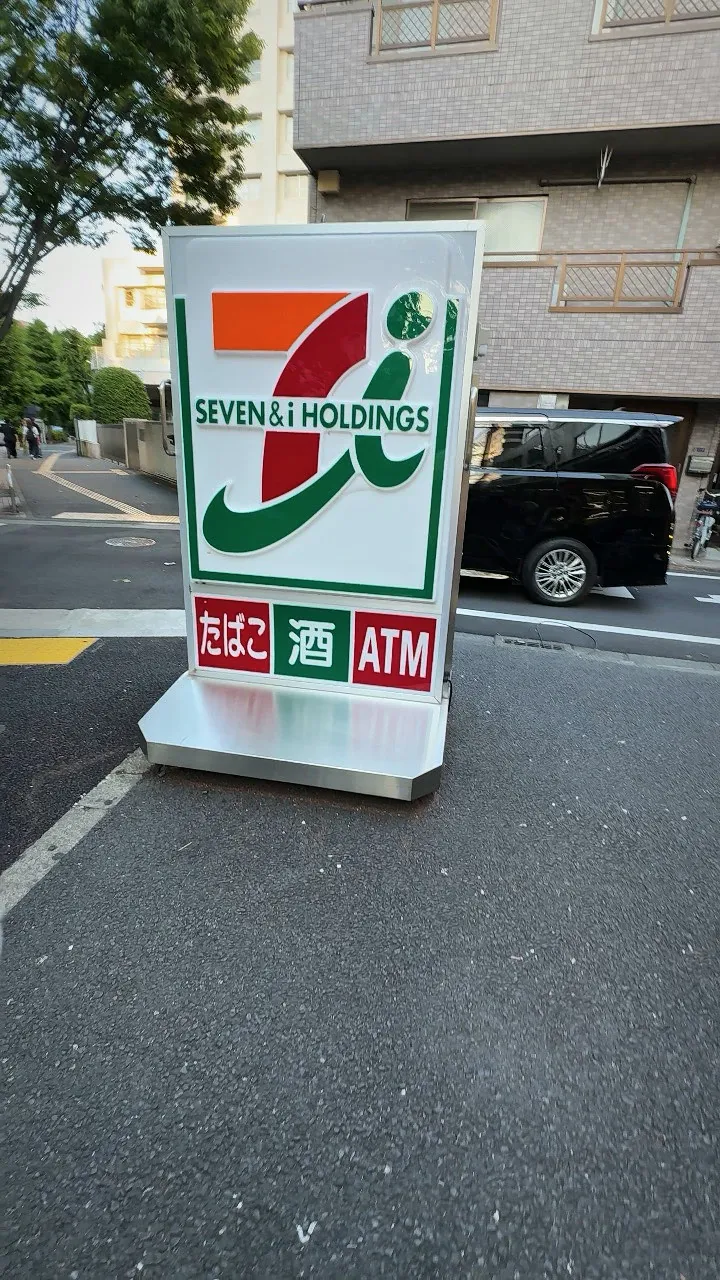 A 7-Eleven sign with an ATM logo standing on the corner of a quiet street in Shinjuku City, Tokyo.