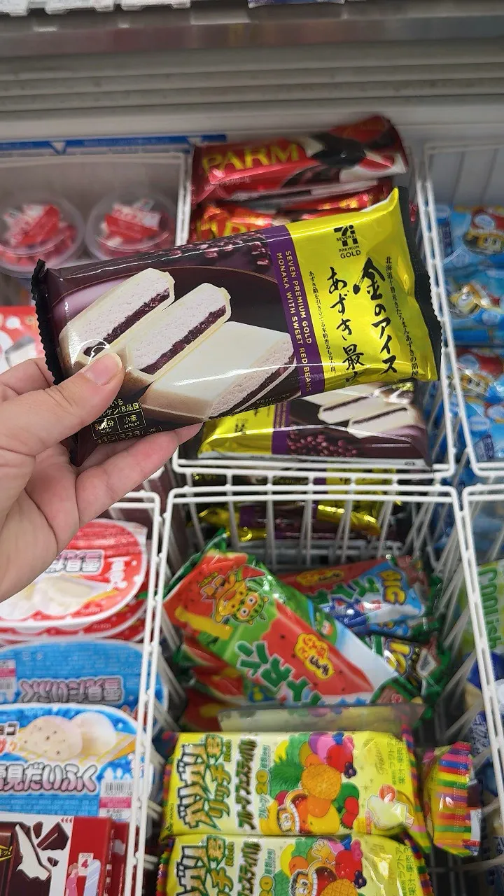 A hand holding a Seven Premium Gold red bean monaka ice cream above a 7-Eleven freezer in Tokyo.