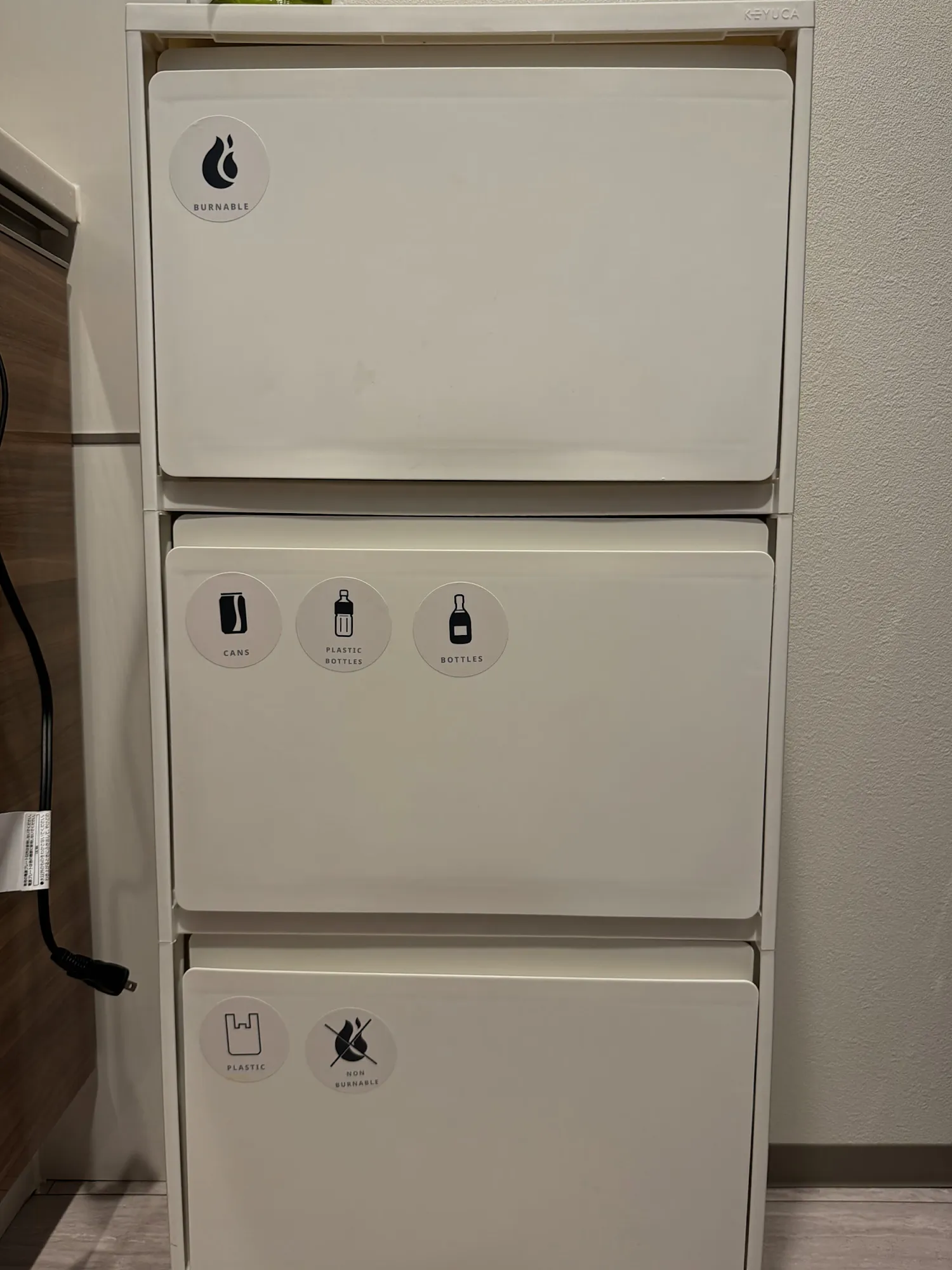 A white three-tier recycling bin in a Tokyo apartment, each drawer labeled with icons for burnable trash, cans, plastic bottles, glass bottles, and non-burnable waste.