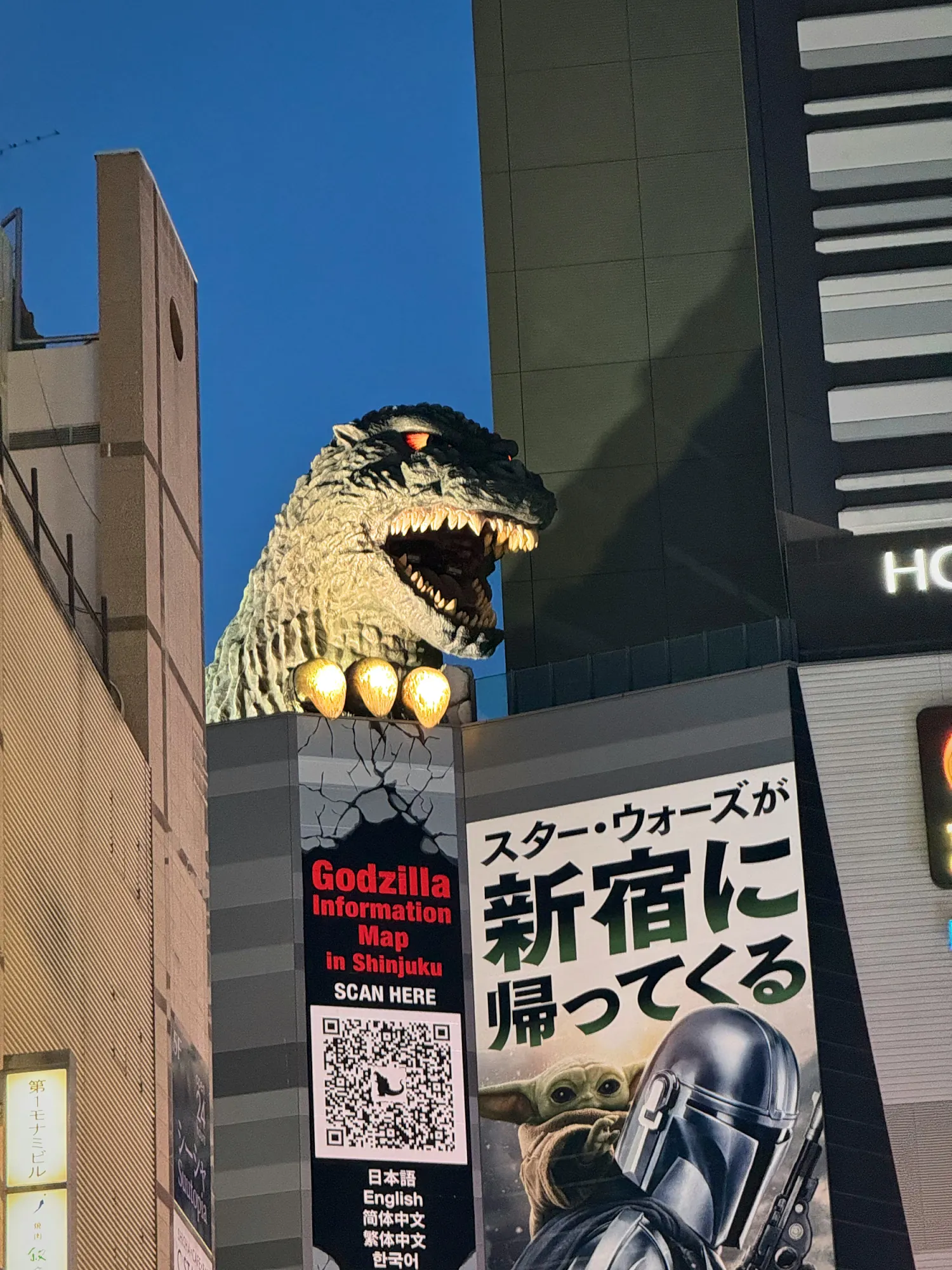 The giant Godzilla head on top of a building in Kabukicho, Shinjuku, at dusk, above a Godzilla sign.