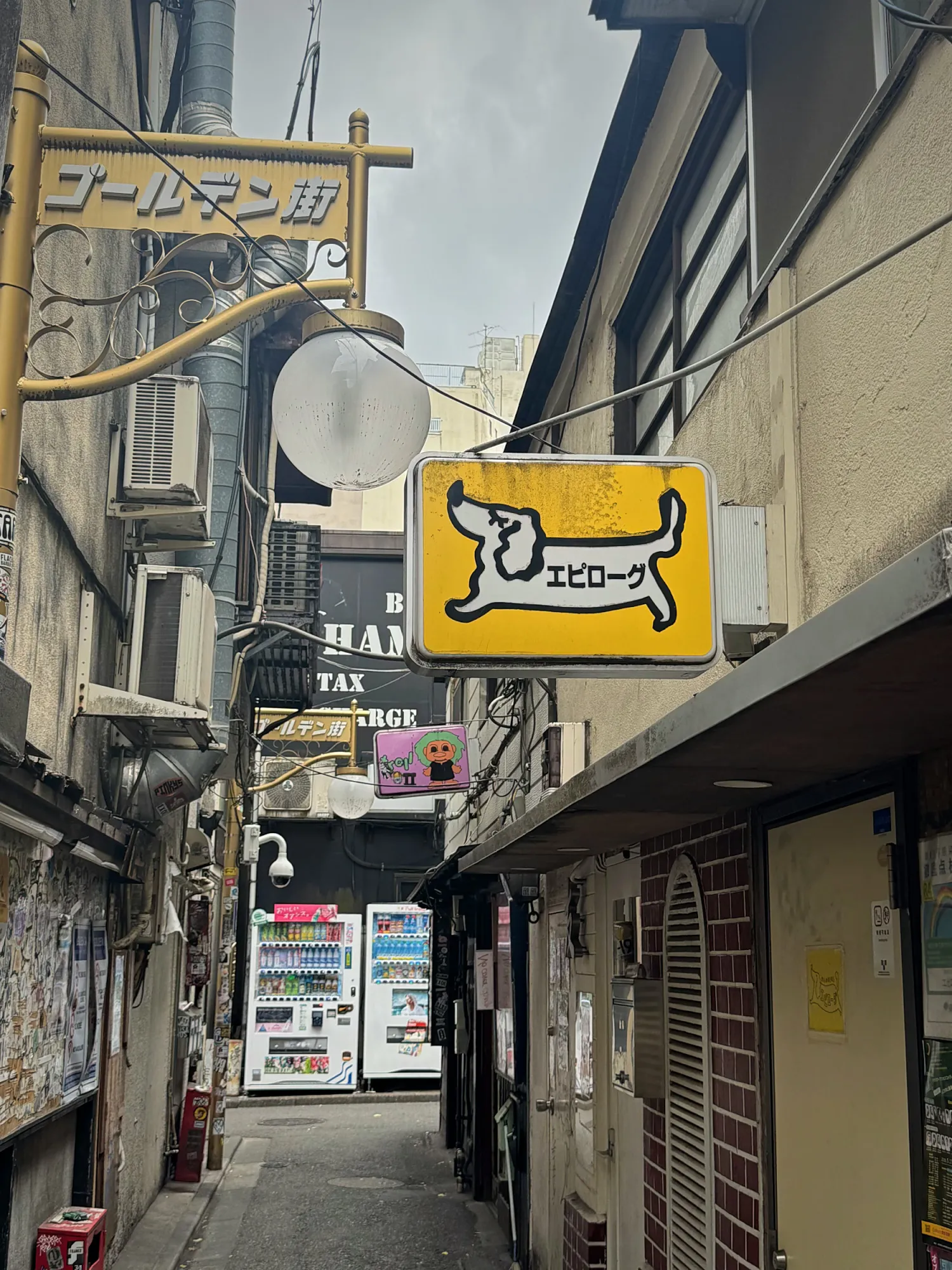 The yellow Golden Gai sign hanging over a narrow alley of small bars in Shinjuku, Tokyo, with vending machines at the far end.