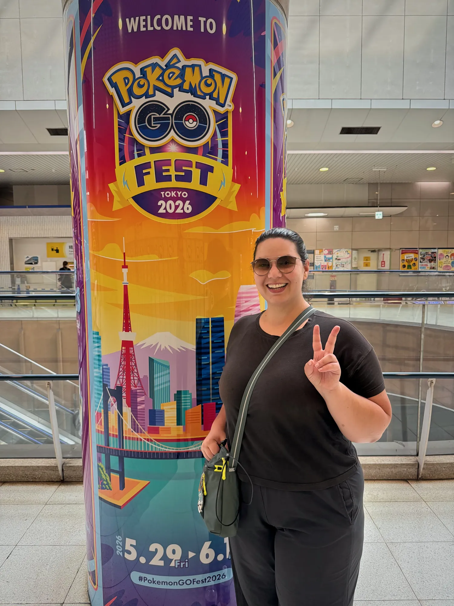 Bernie making a peace sign next to a tall Pokémon GO Fest Tokyo pillar showing the 5.29 to 6.1 dates inside a train station.