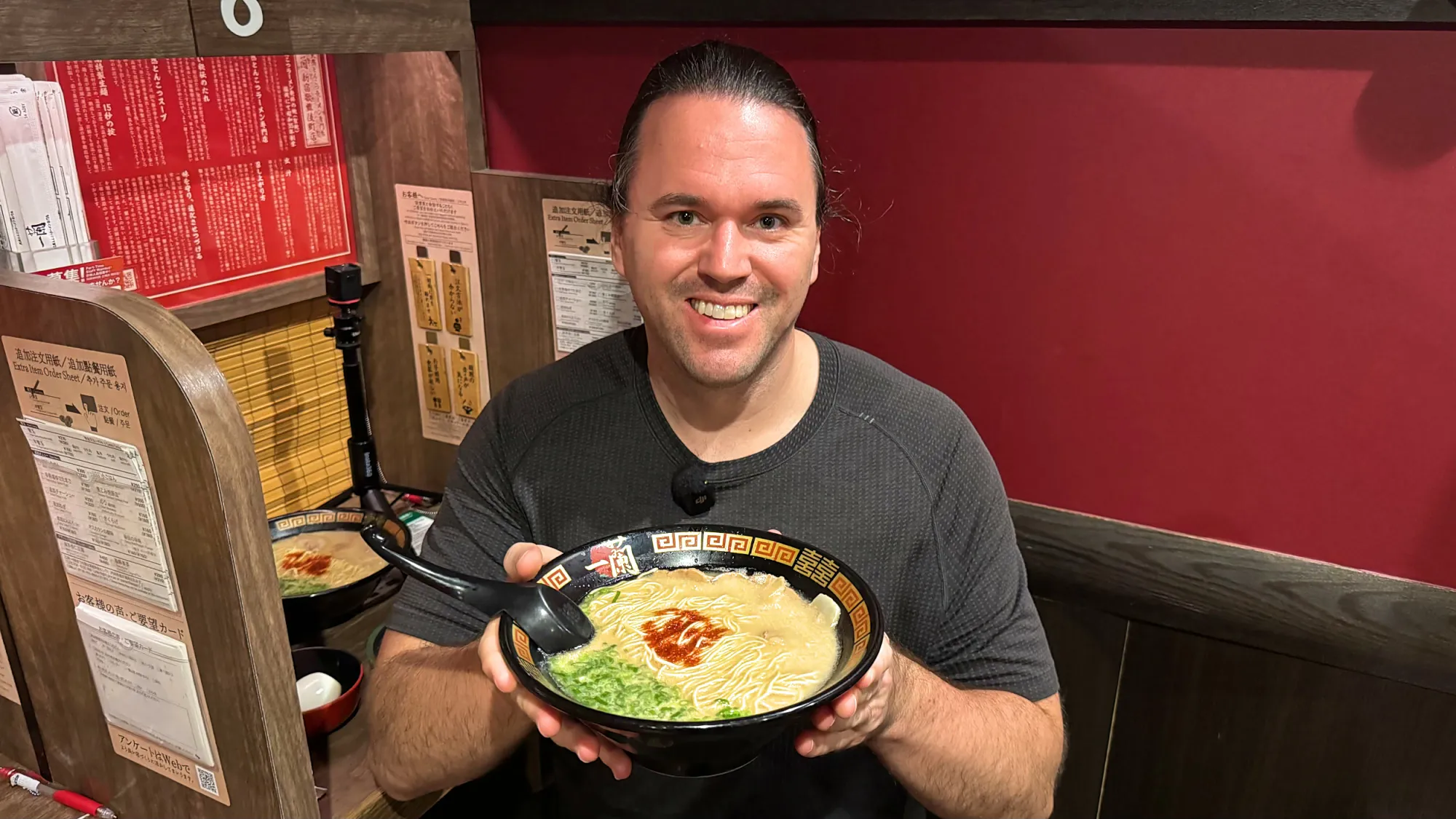 Cream smiling and holding up his bowl of Ichiran tonkotsu ramen in a solo booth.
