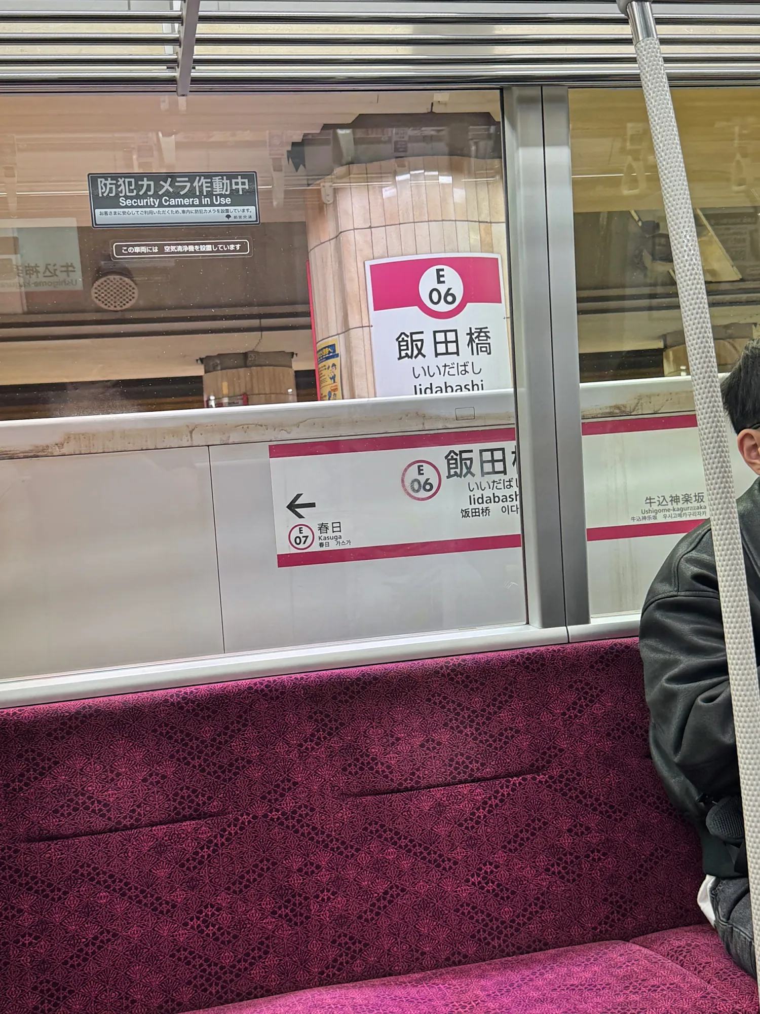 The Iidabashi station sign, E06, seen through the window of an Oedo Line train above purple seats.