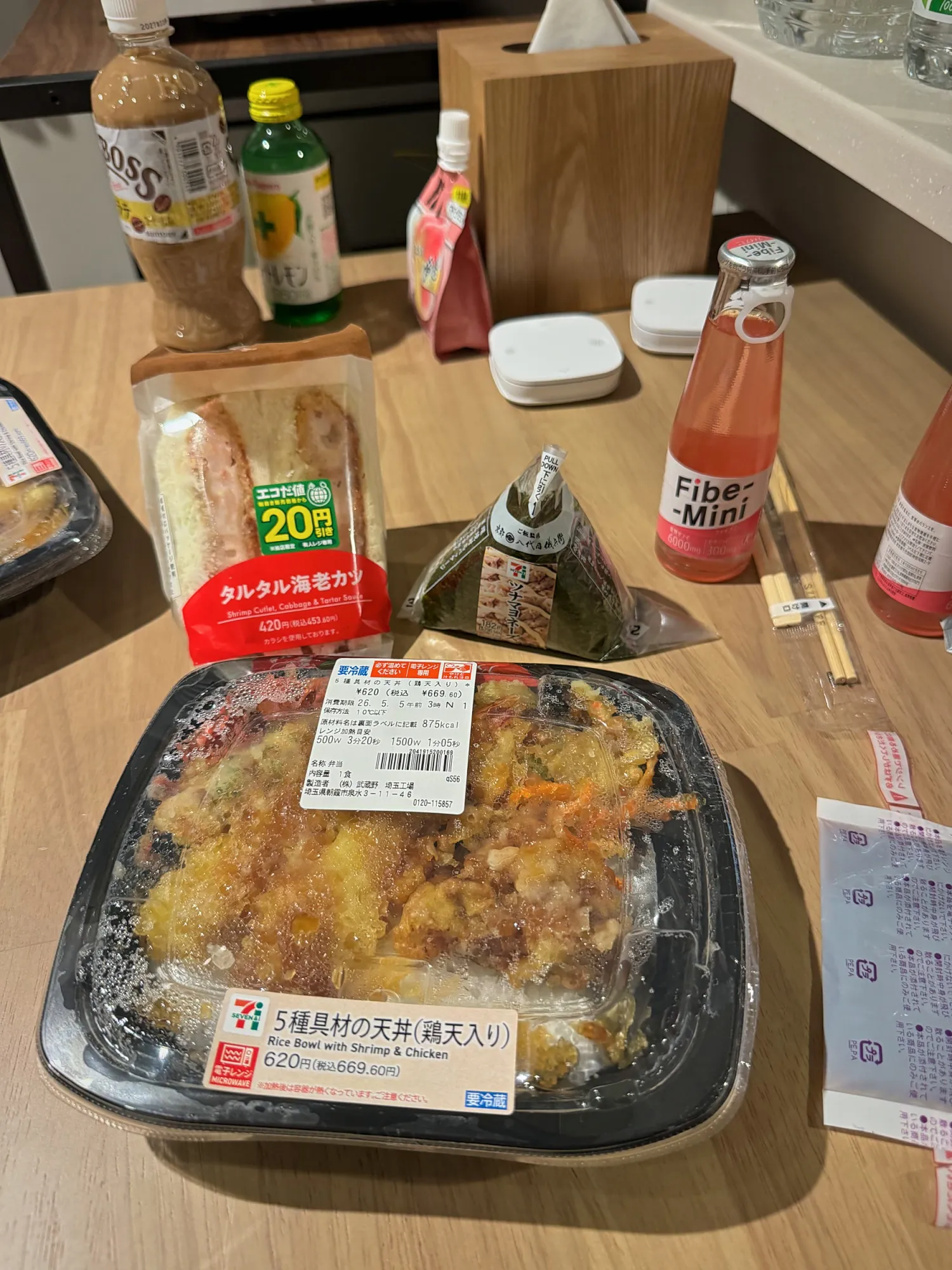 A 7-Eleven dinner spread on a table in Tokyo, a tempura rice bowl, an onigiri, a sandwich, and drinks