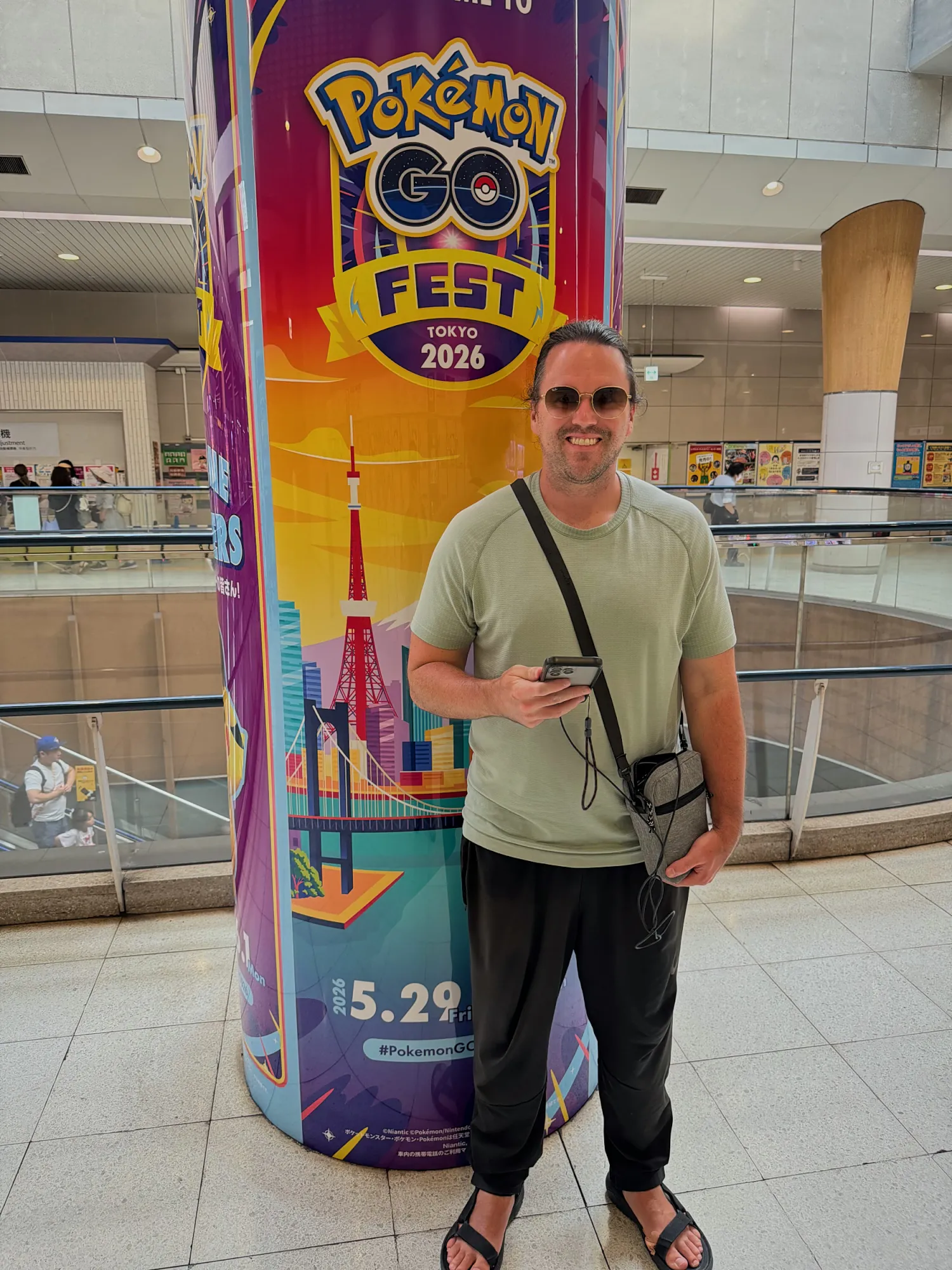 Cream holding up his phone with Pokémon GO open next to a colorful Pokémon GO Fest Tokyo pillar in a mall.