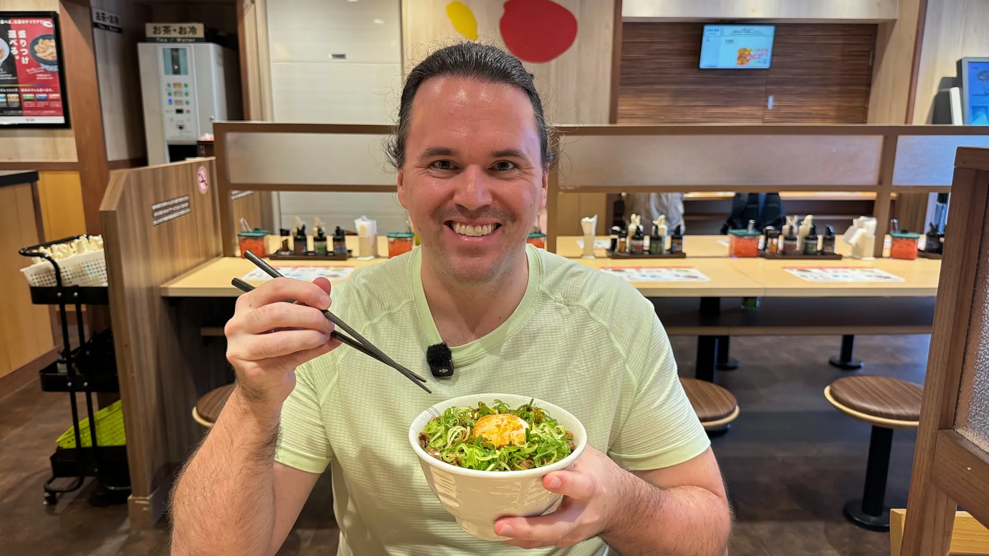 Cream lifting gyudon with chopsticks from a bowl topped with green onions and egg in a Matsuya booth