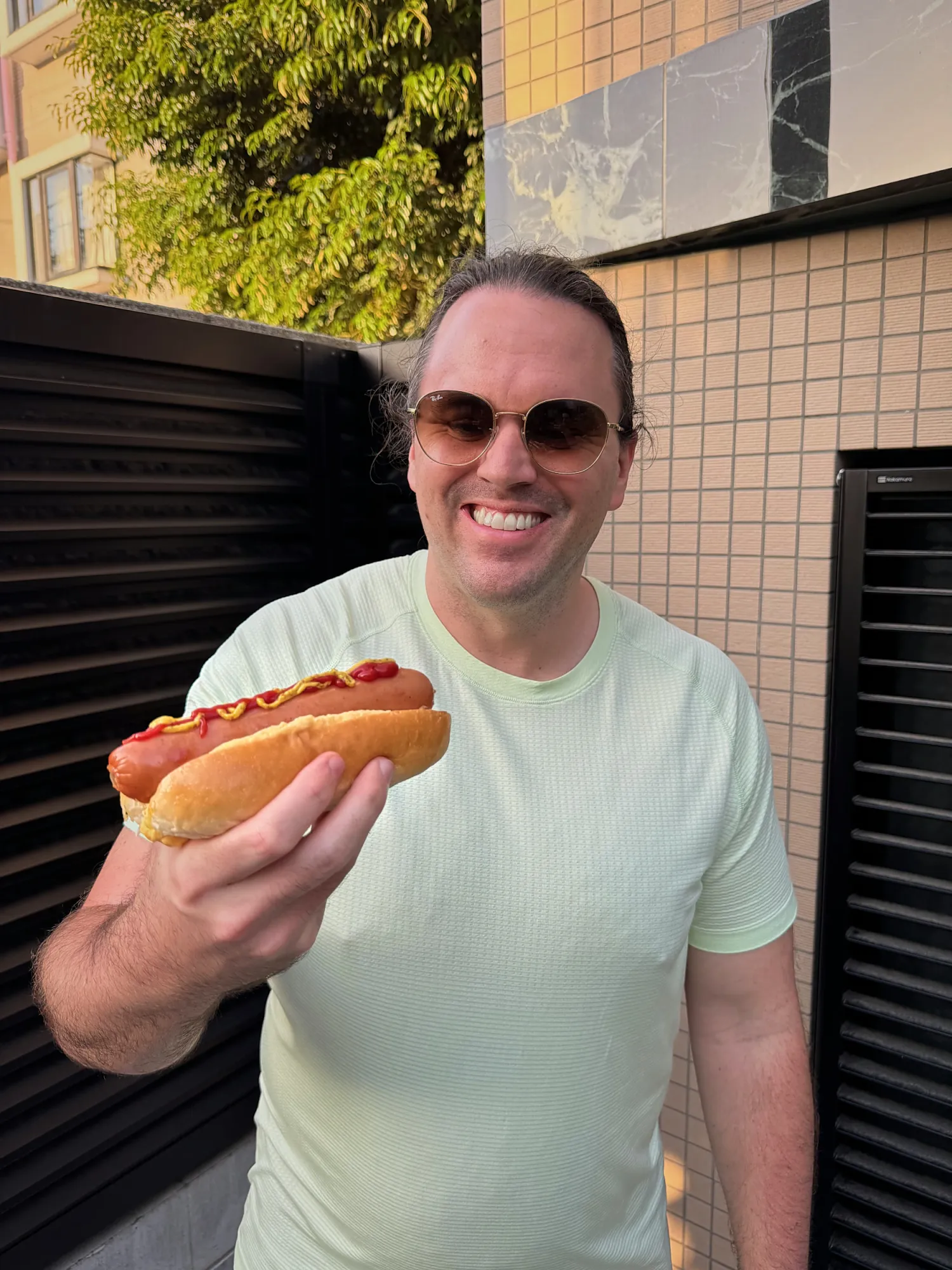 Cream grinning and holding up the 7-Eleven hot dog hack, a Big Frank sausage in a bun with ketchup and mustard
