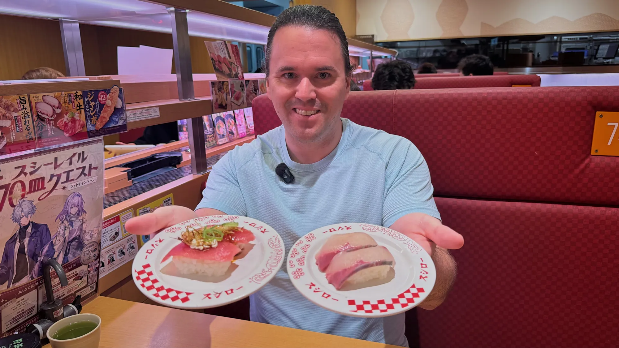Cream smiling in a red Sushiro booth in Tokyo, holding up two plates of nigiri.