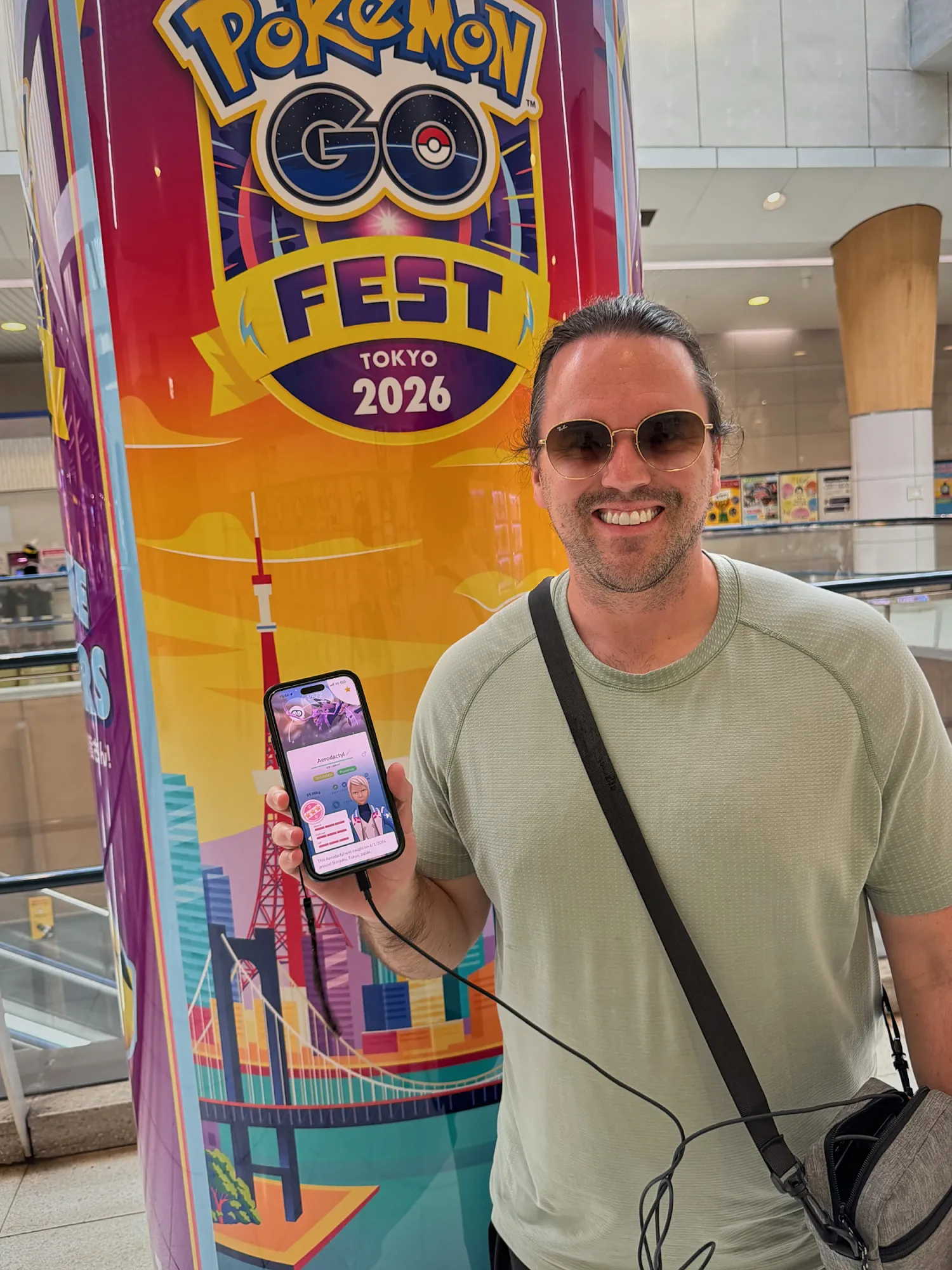 Cream smiling and holding up his phone with Pokémon GO open beside a tall Pokémon GO Fest Tokyo 2026 pillar.
