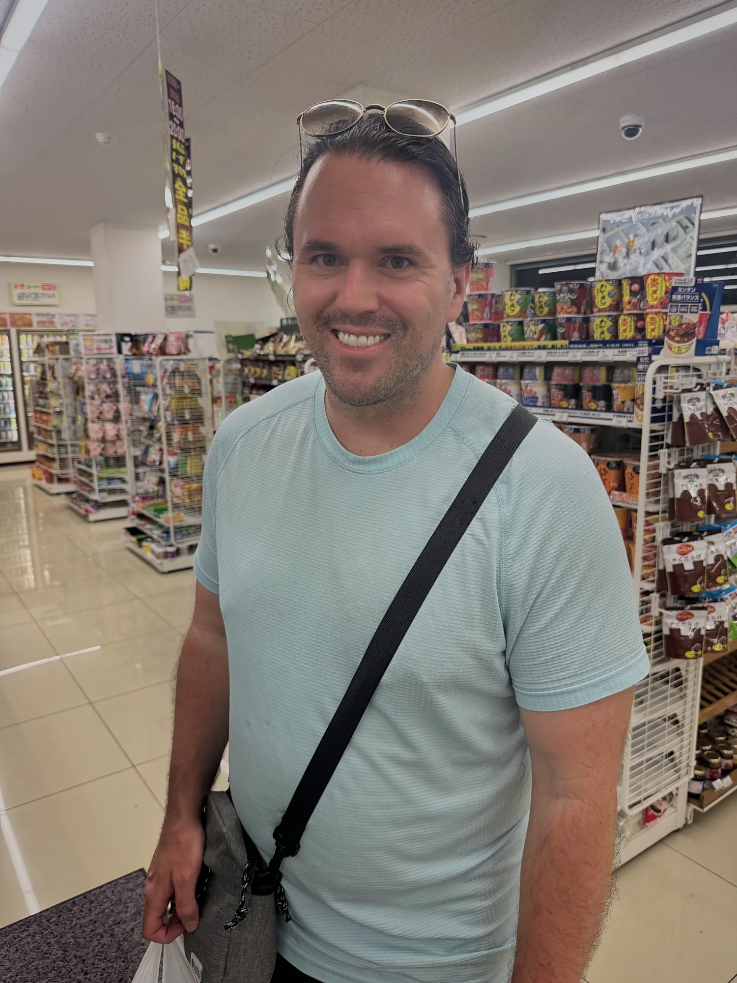 A smiling traveler standing in the aisle of a Japanese convenience store lined with snacks and drinks.