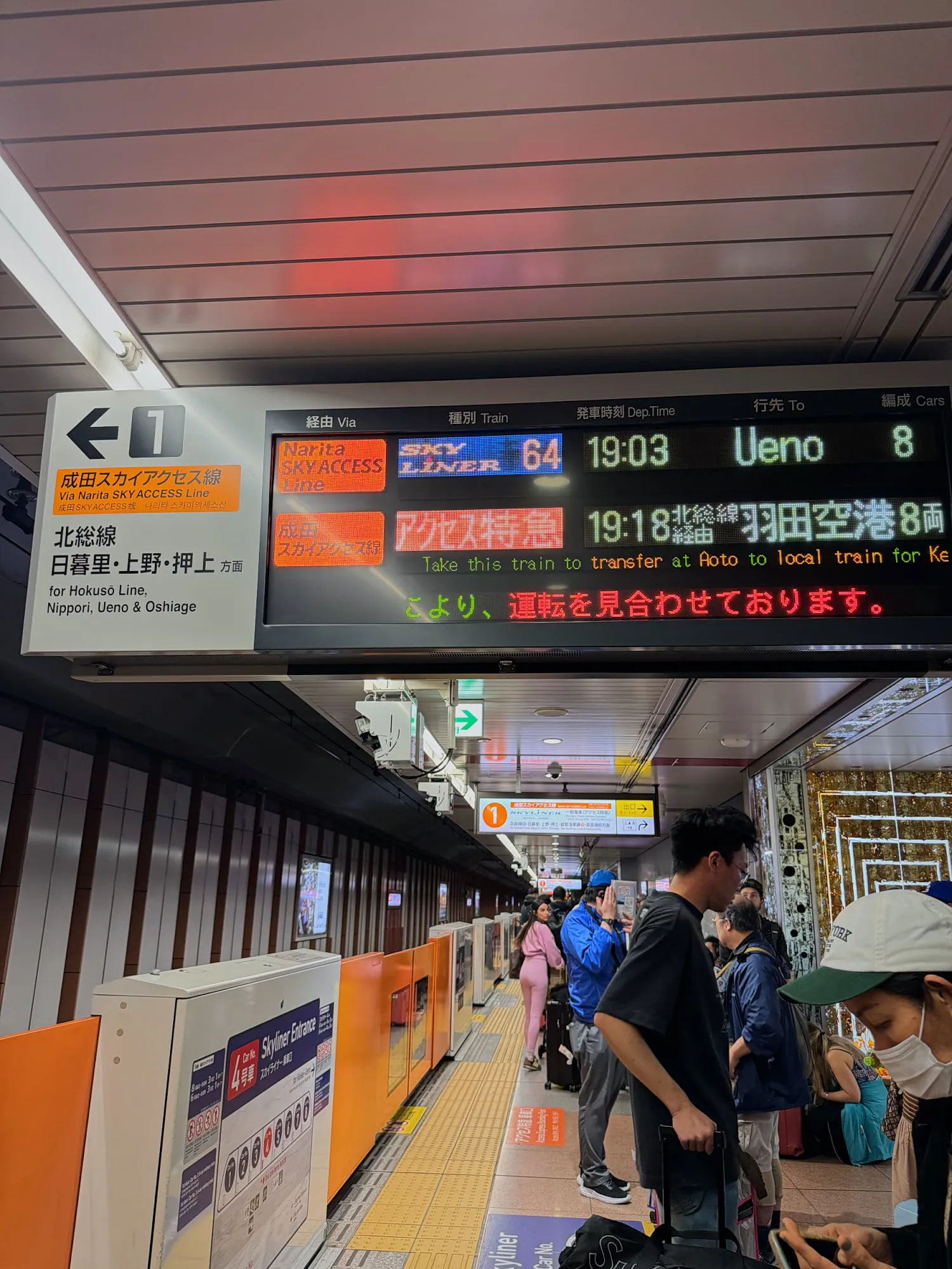 A digital departure board at a Narita Airport train platform showing a Skyliner to Ueno, with travelers waiting below.