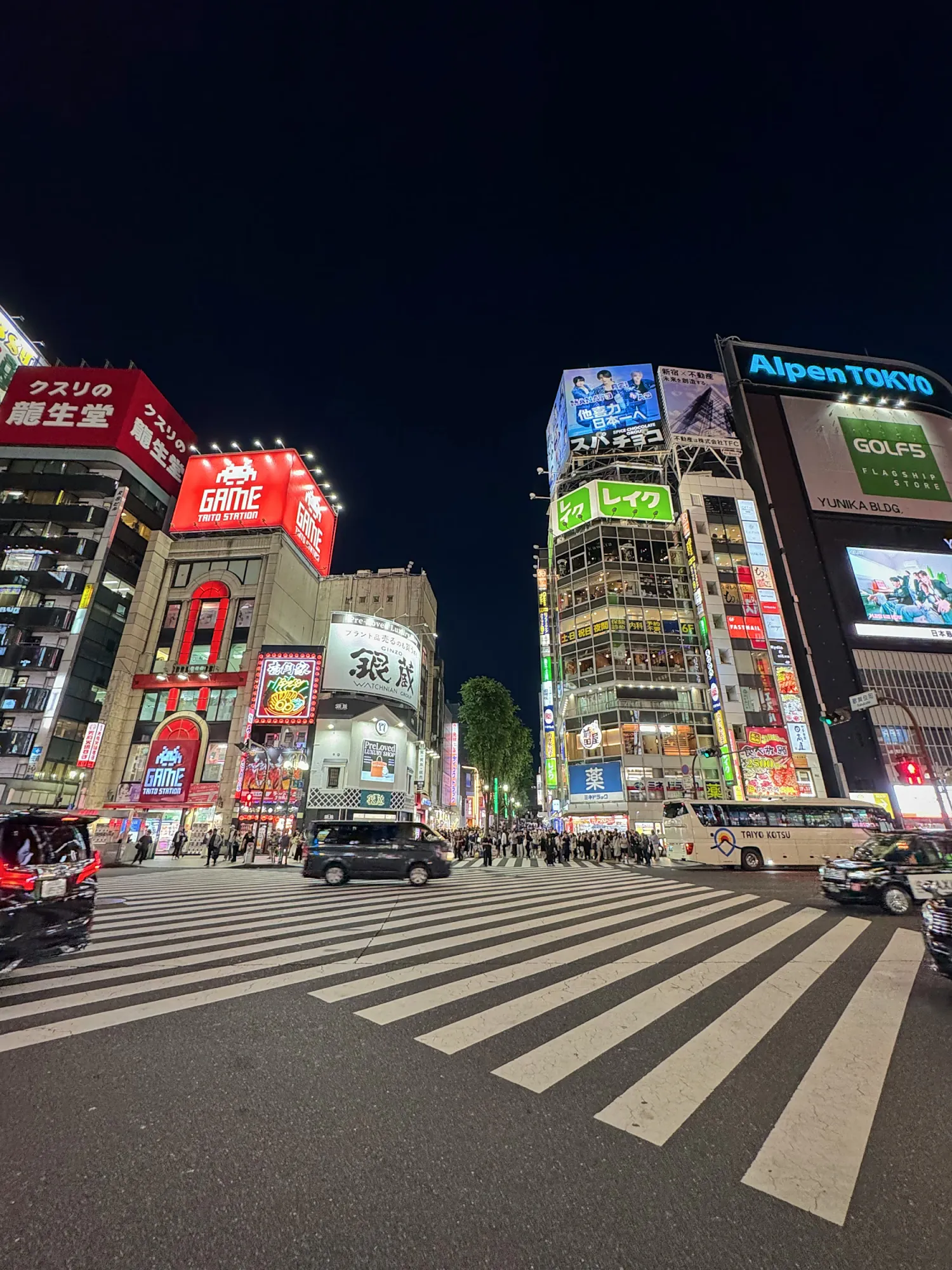 A wide Shinjuku intersection at night, lit by neon signs and billboards on the buildings around it, Tokyo.