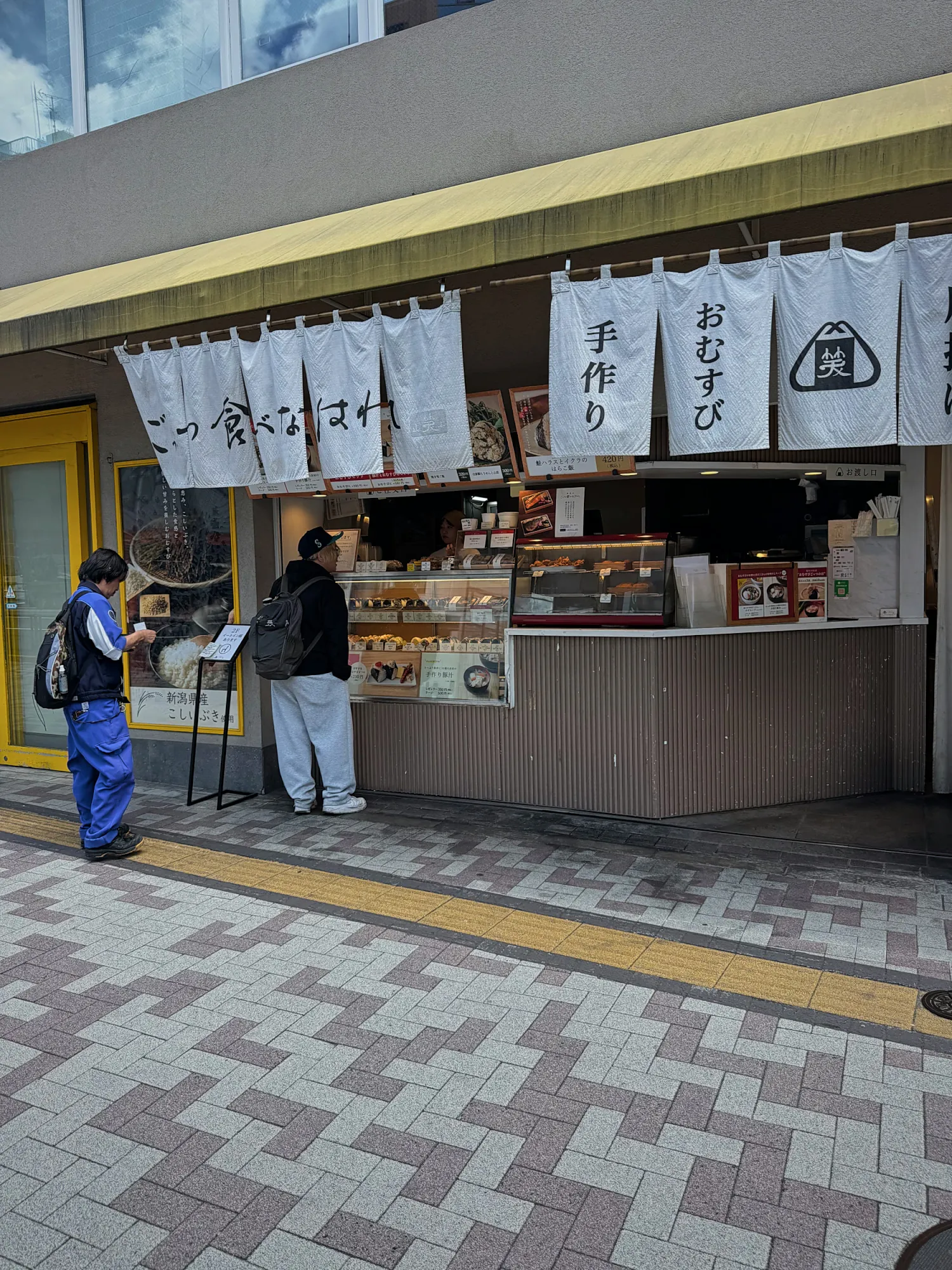 Customers ordering at a small onigiri shop with white noren curtains under a yellow awning in Nakameguro, Tokyo.