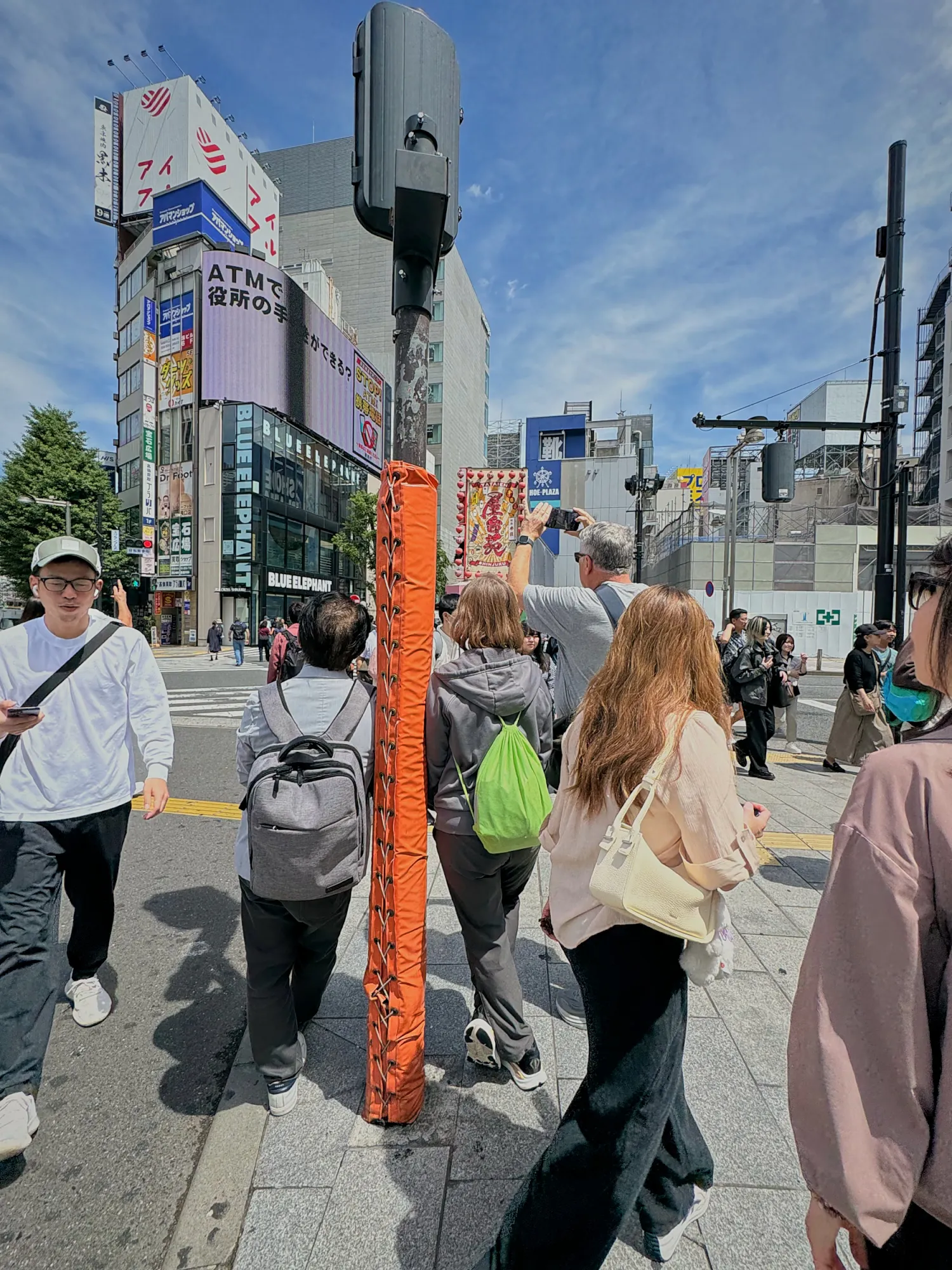 People crossing a wide street in Shinjuku, Tokyo, past shops, signs, and a tall orange pole.