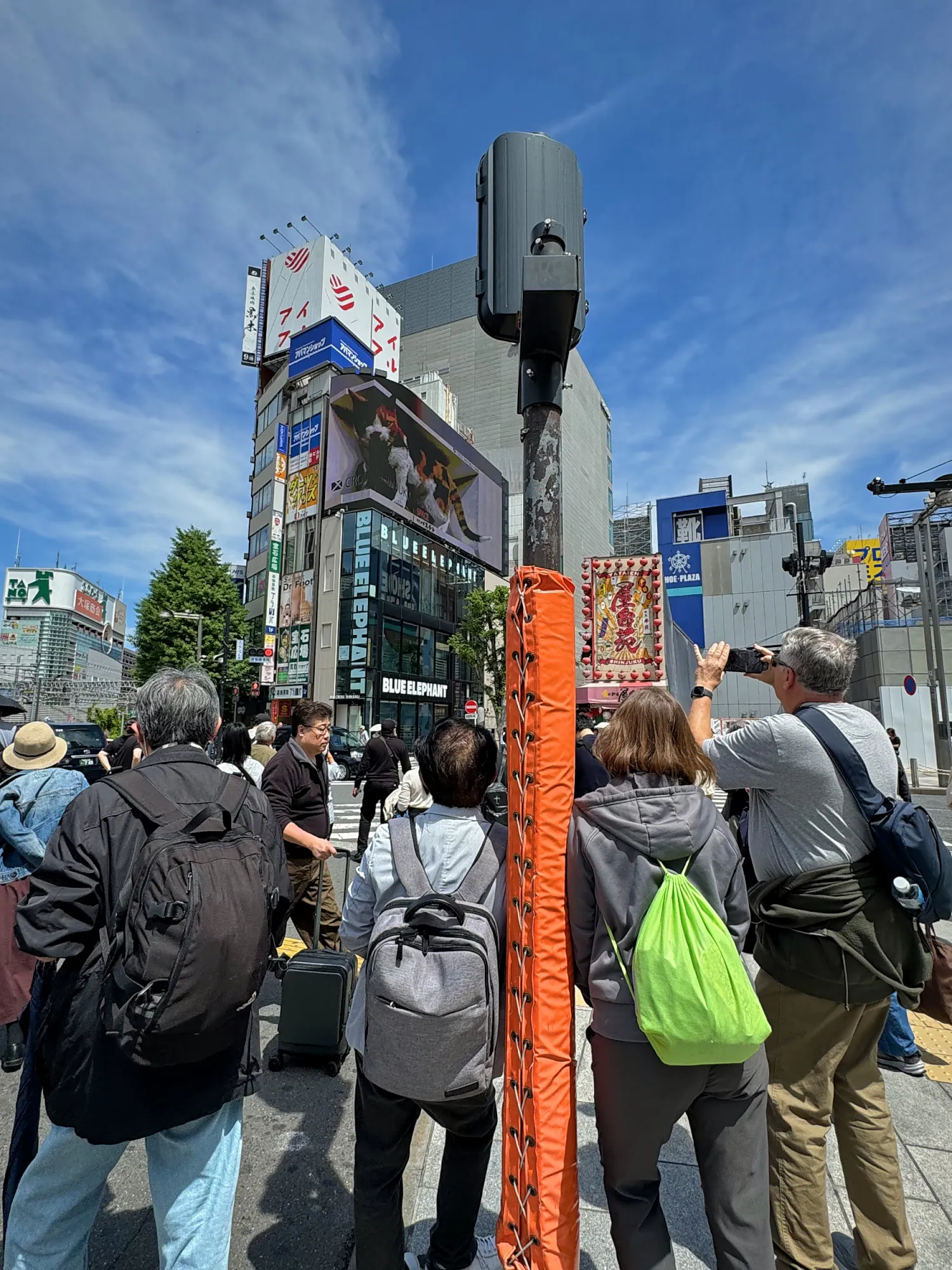 A crowd of people with backpacks waiting at a crosswalk in Shinjuku, Tokyo, on a sunny day.