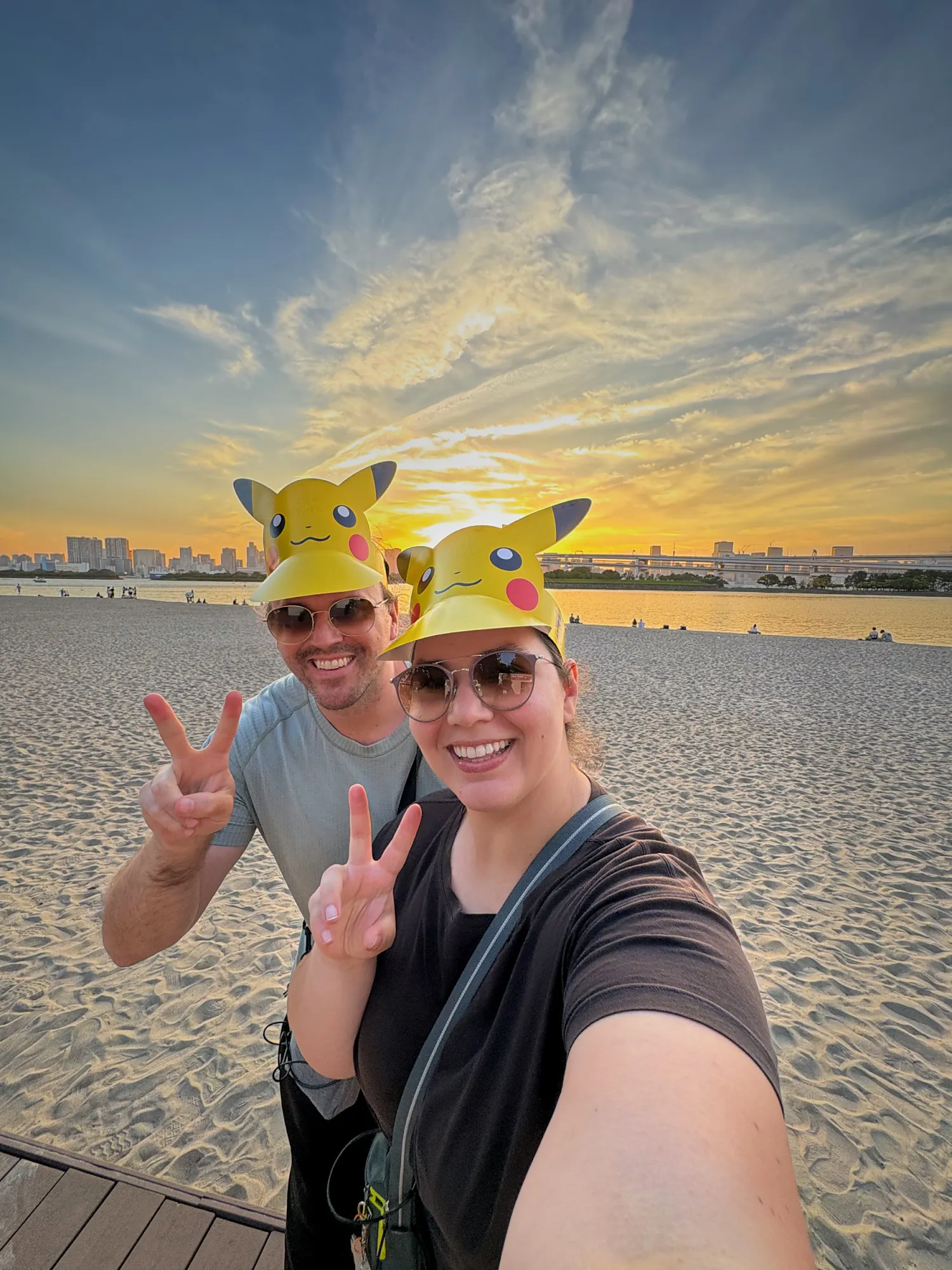 Bernie and Cream in Pikachu visors taking a selfie on Odaiba beach at sunset.